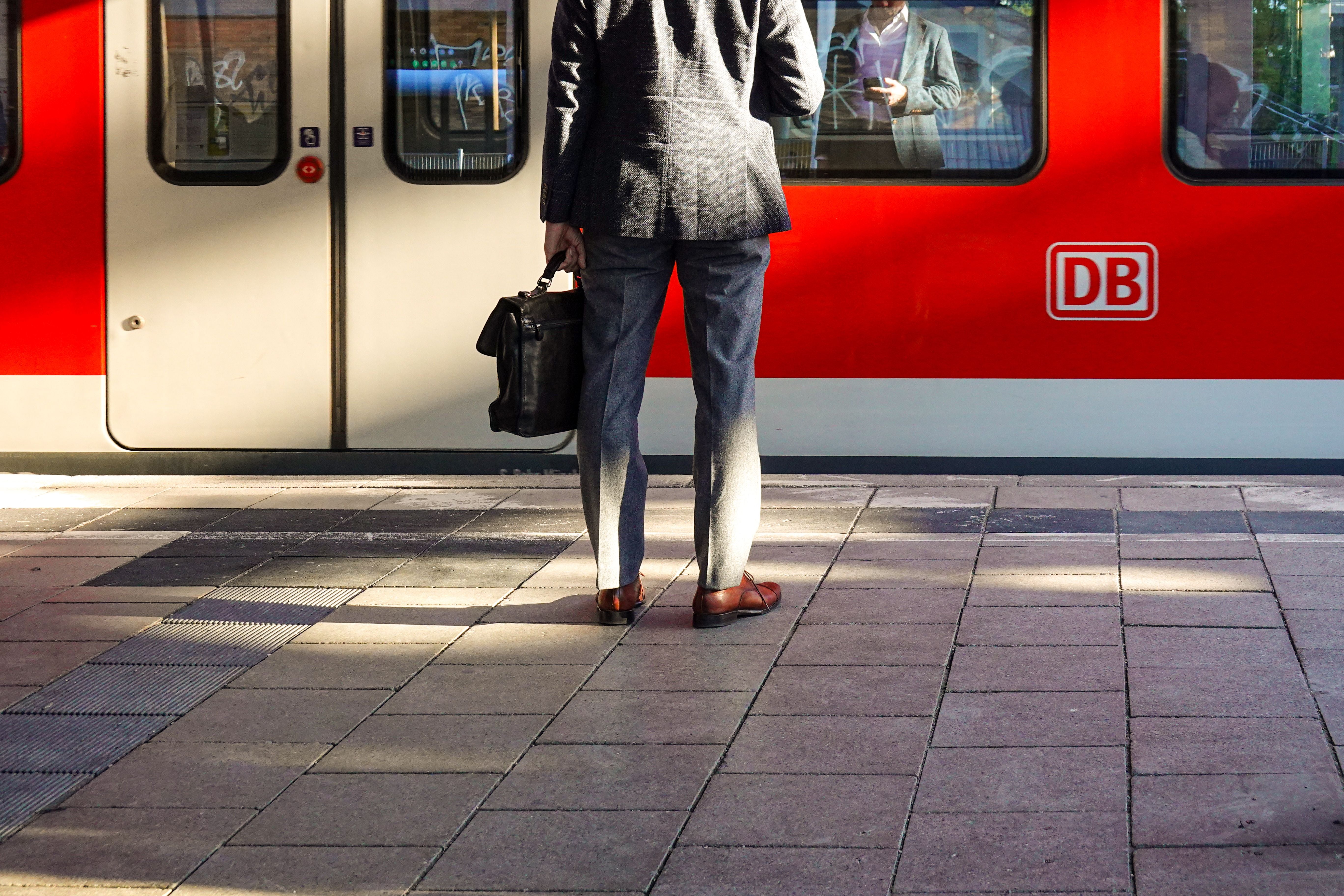 Ein Mann im Anzug mit Aktentasche steht am Bahnsteig vor einem roten DB-Zug.