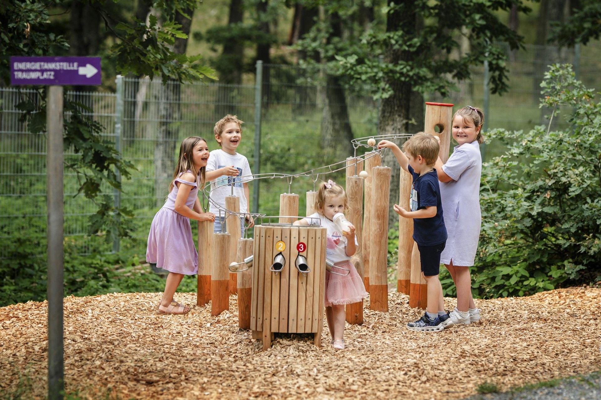 Fünf Kinder spielen an einer Kugelbahn aus Holz im Freien vor einem Zaun und Bäumen.