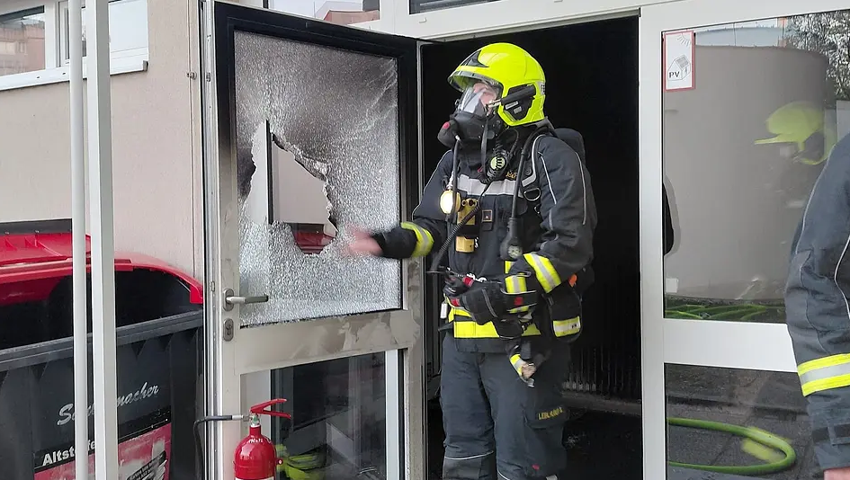 Feuerwehrmann mit Atemschutzmaske vor eingeschlagener Glastür nach einem Brand.