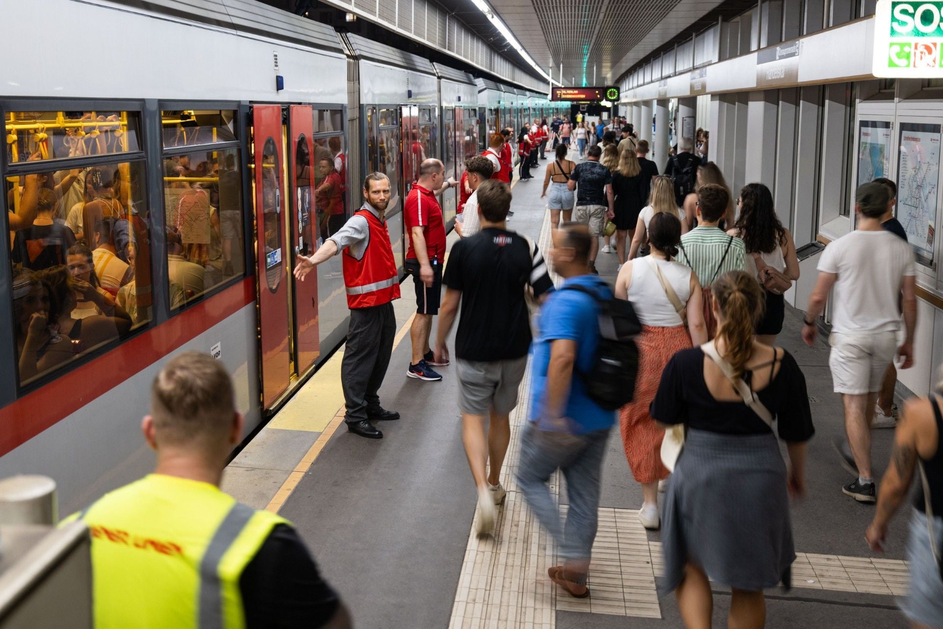 Viele Menschen steigen an einem U-Bahnsteig ein und aus, Ordner tragen rote Westen.