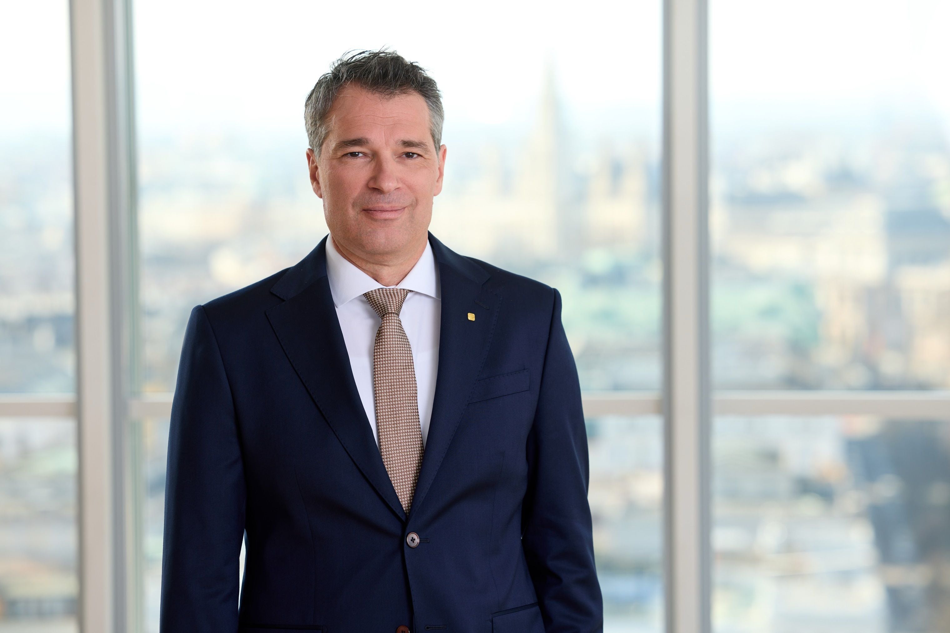 Ein Mann im Anzug steht vor einem großen Fenster mit unscharfem Stadtblick im Hintergrund.