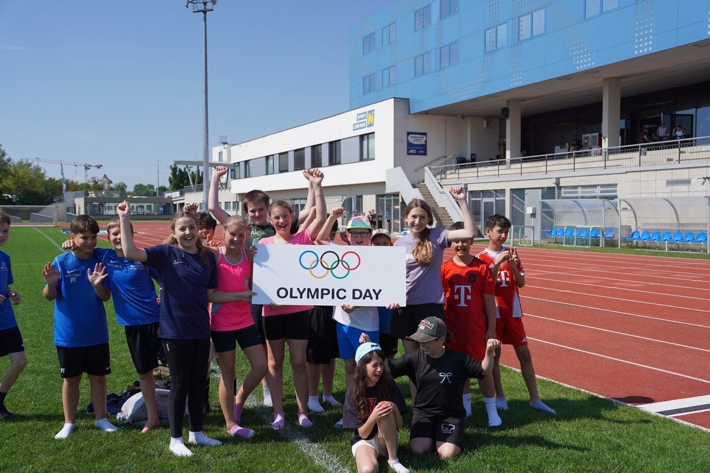 Eine Kindergruppe hält ein „Olympic Day“-Schild auf einem Sportplatz vor dem Olympiazentrum.