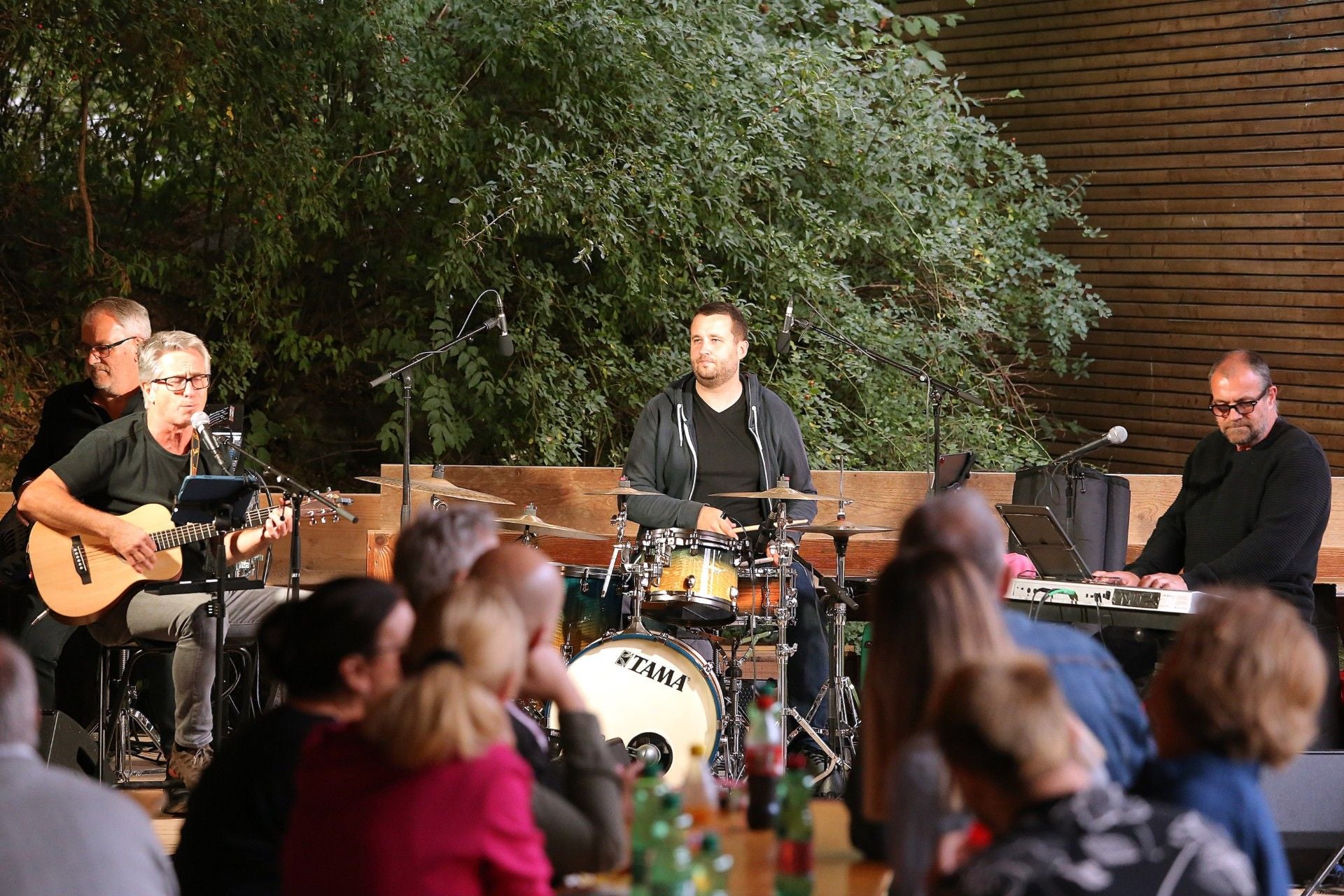 Eine Band spielt live auf einer Bühne, vor Publikum mit Getränken an Tischen.