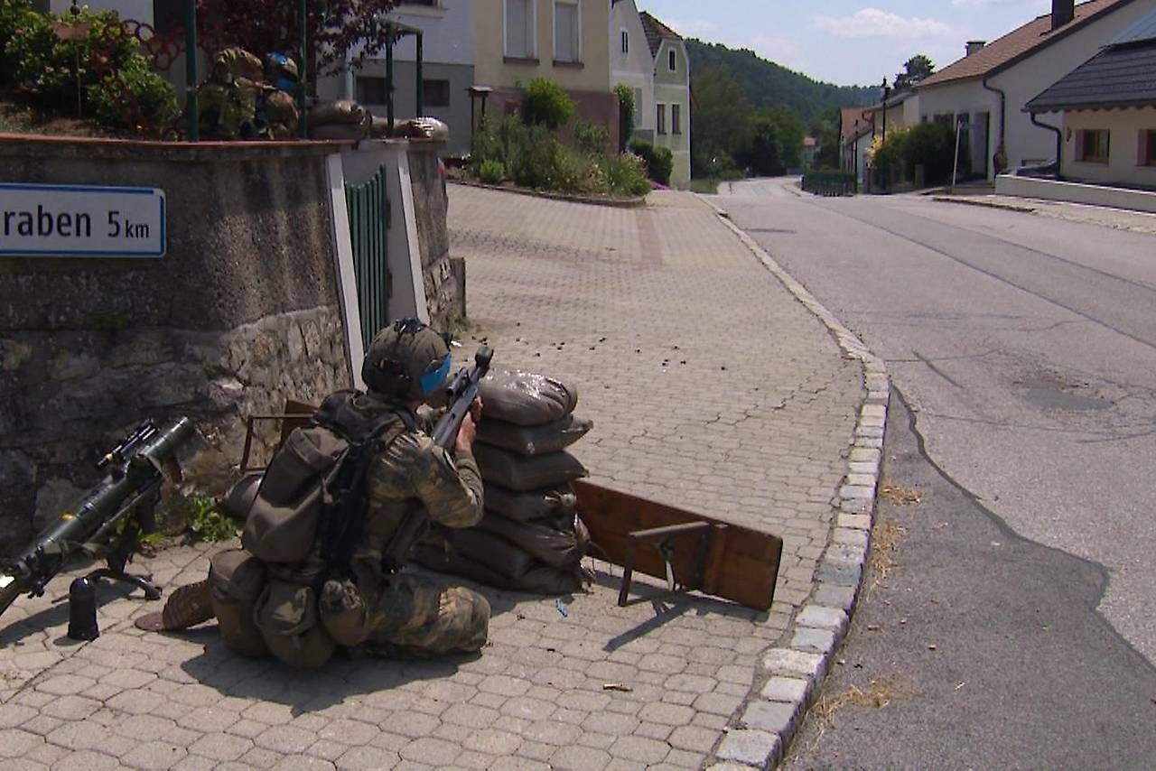 Ein Soldat kauert hinter Sandsäcken und zielt mit seinem Gewehr eine leere Straße entlang.