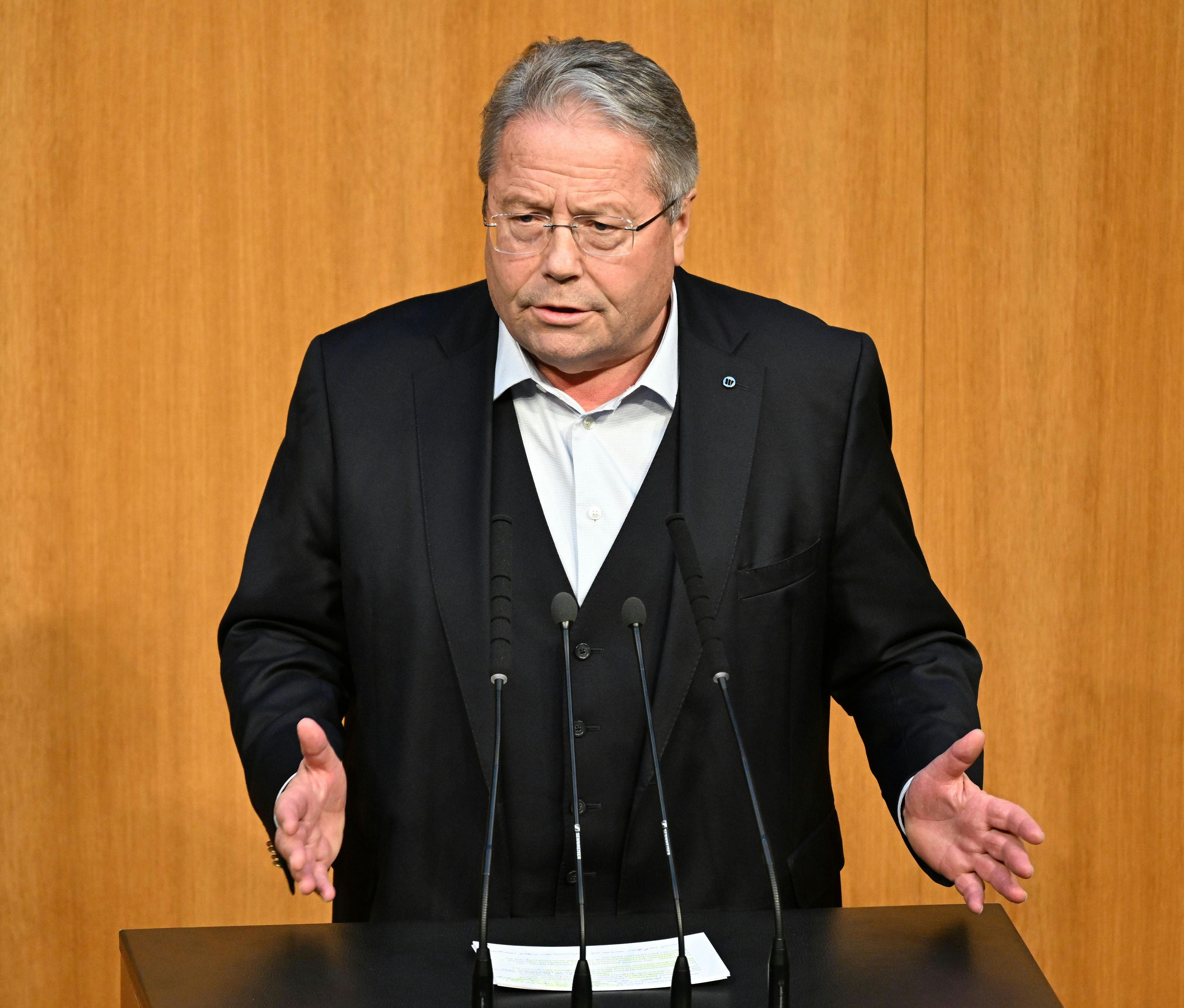 Ein Mann im Anzug spricht gestikulierend an einem Rednerpult mit Mikrofonen im Parlament.