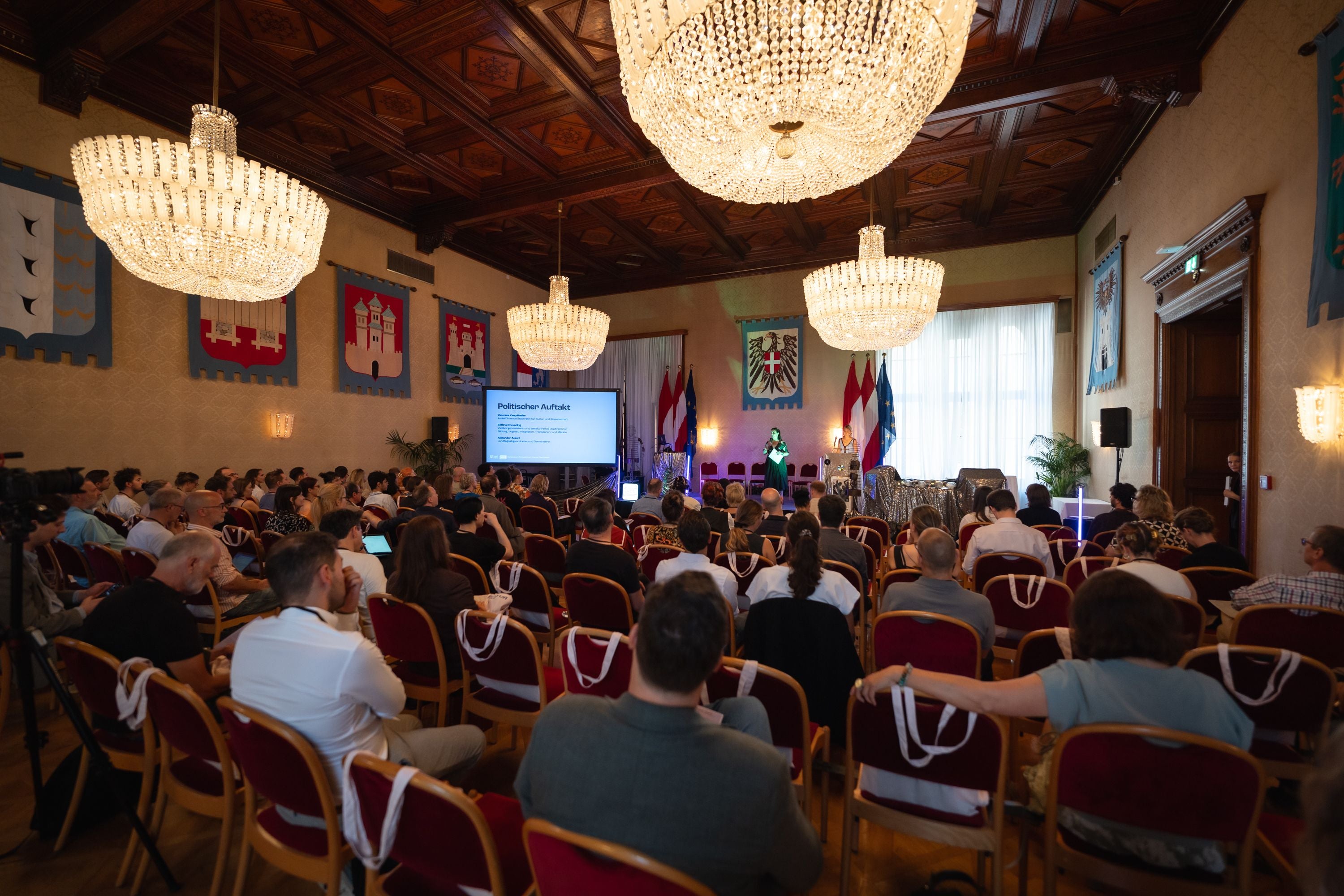 Publikum sitzt im prunkvollen Saal des Wiener Rathauses, Vortrag mit Präsentation vorne.