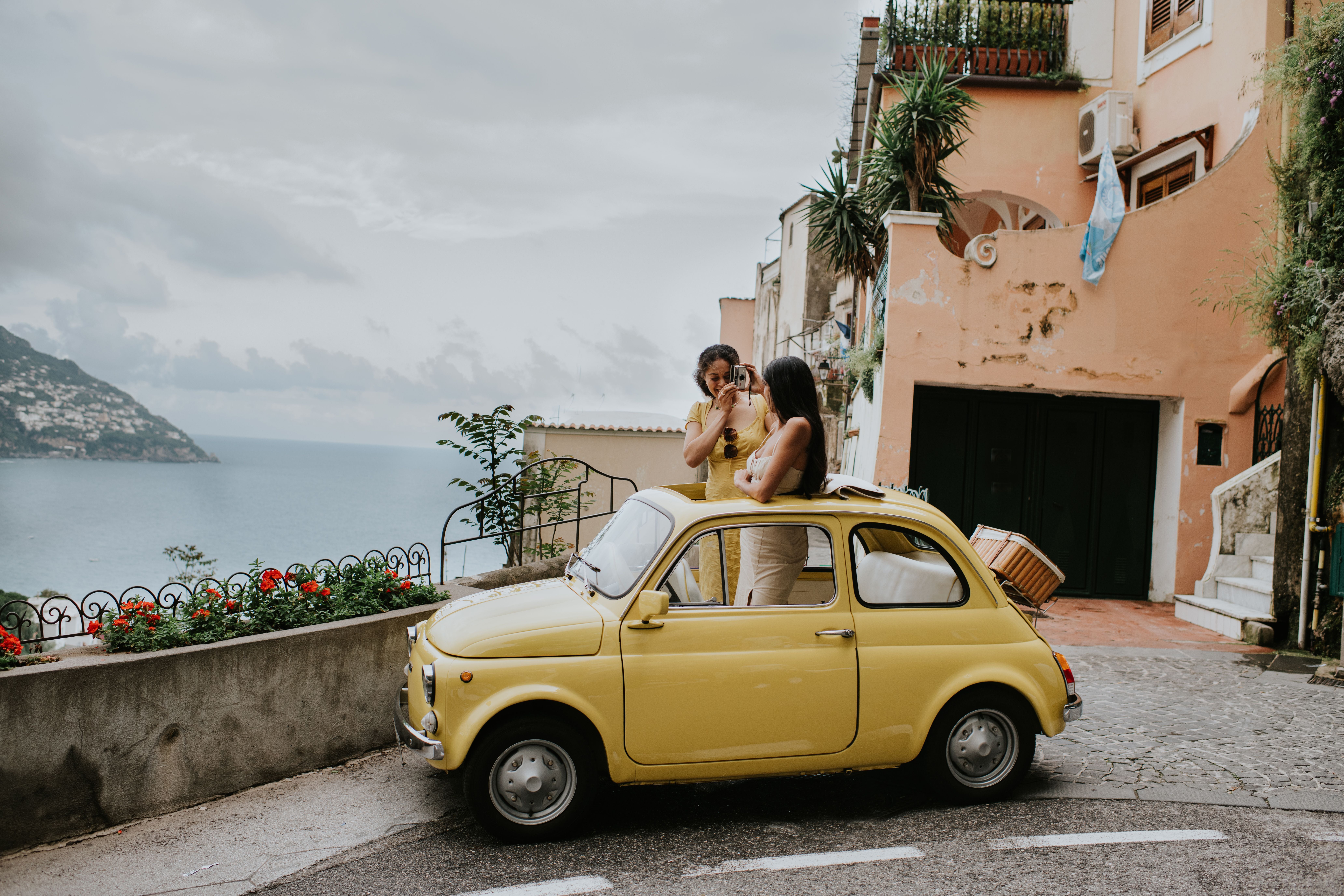Two beautiful young woman inside a small, vintage yellow car. The top remains open. They stand up in the vehicle, through the sunroof and take photos of each other. We can see Italy's famous view of Positano in the background. Depicts a scene of experiential travel, suggesting renting a car, travel tours, European road trips and nostalgia.