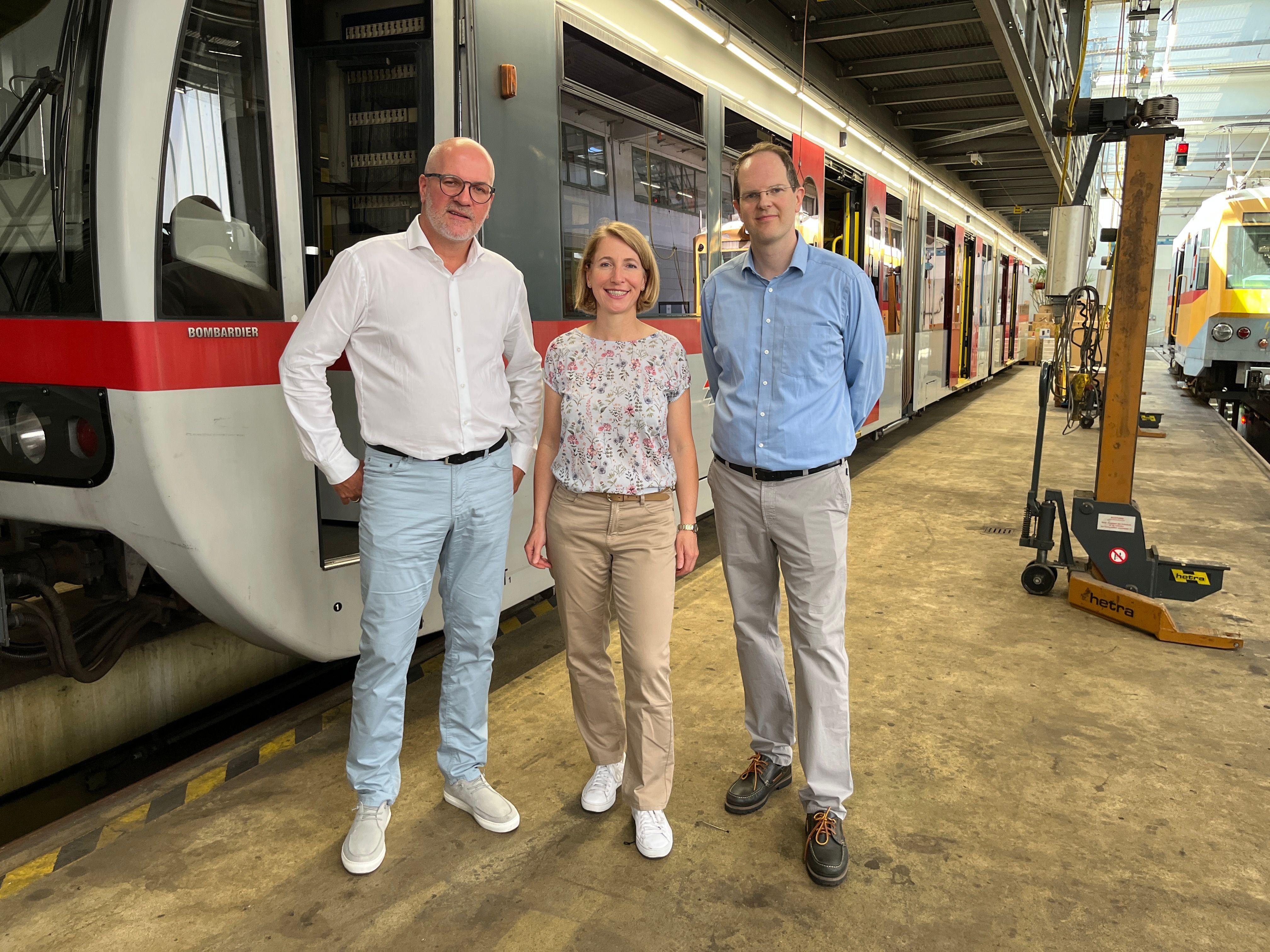 Drei Personen stehen in einer Straßenbahn-Werkstatt neben einer roten Straßenbahn.