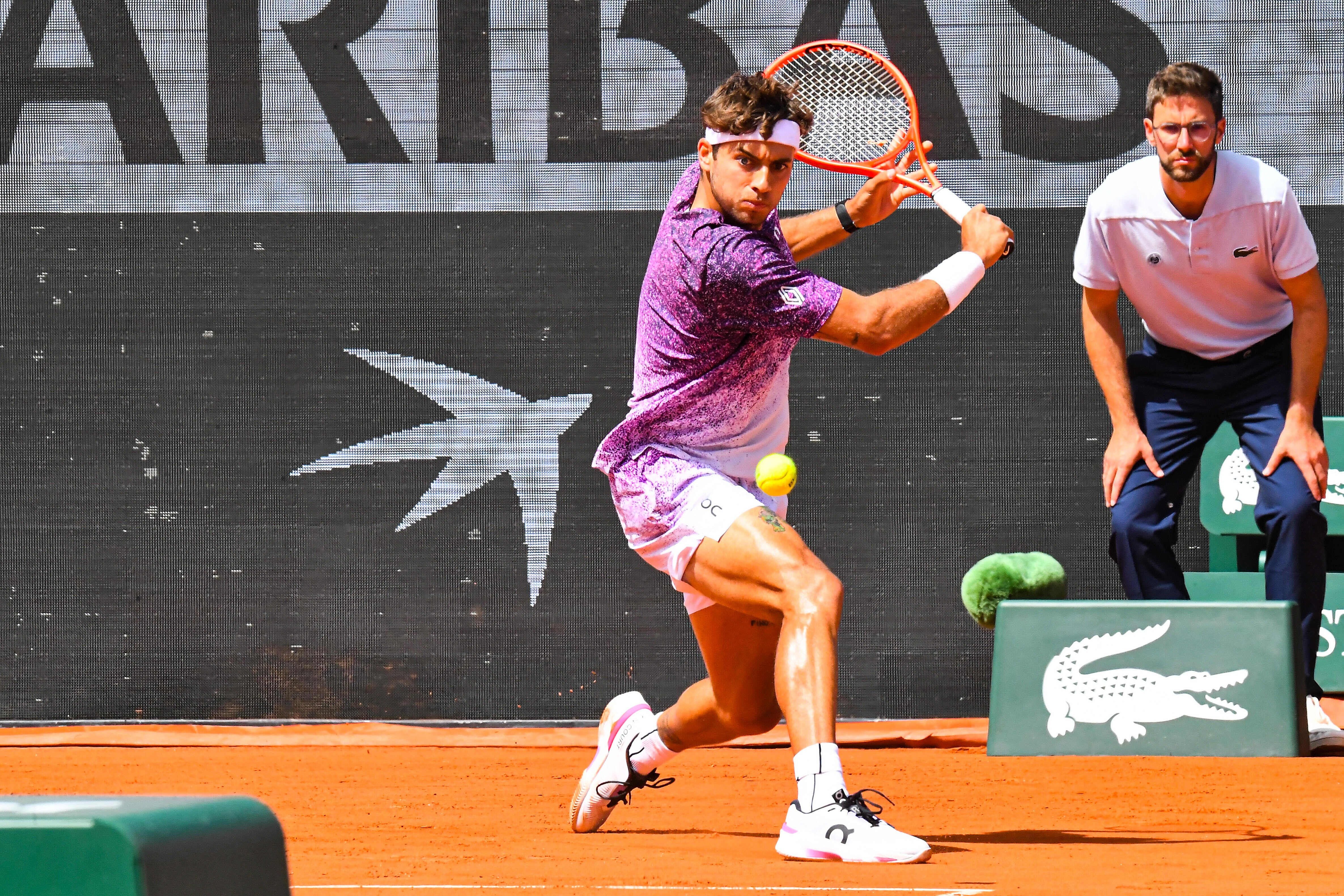Tennisspieler beim Schlag auf Sandplatz, Schiedsrichter im Hintergrund.