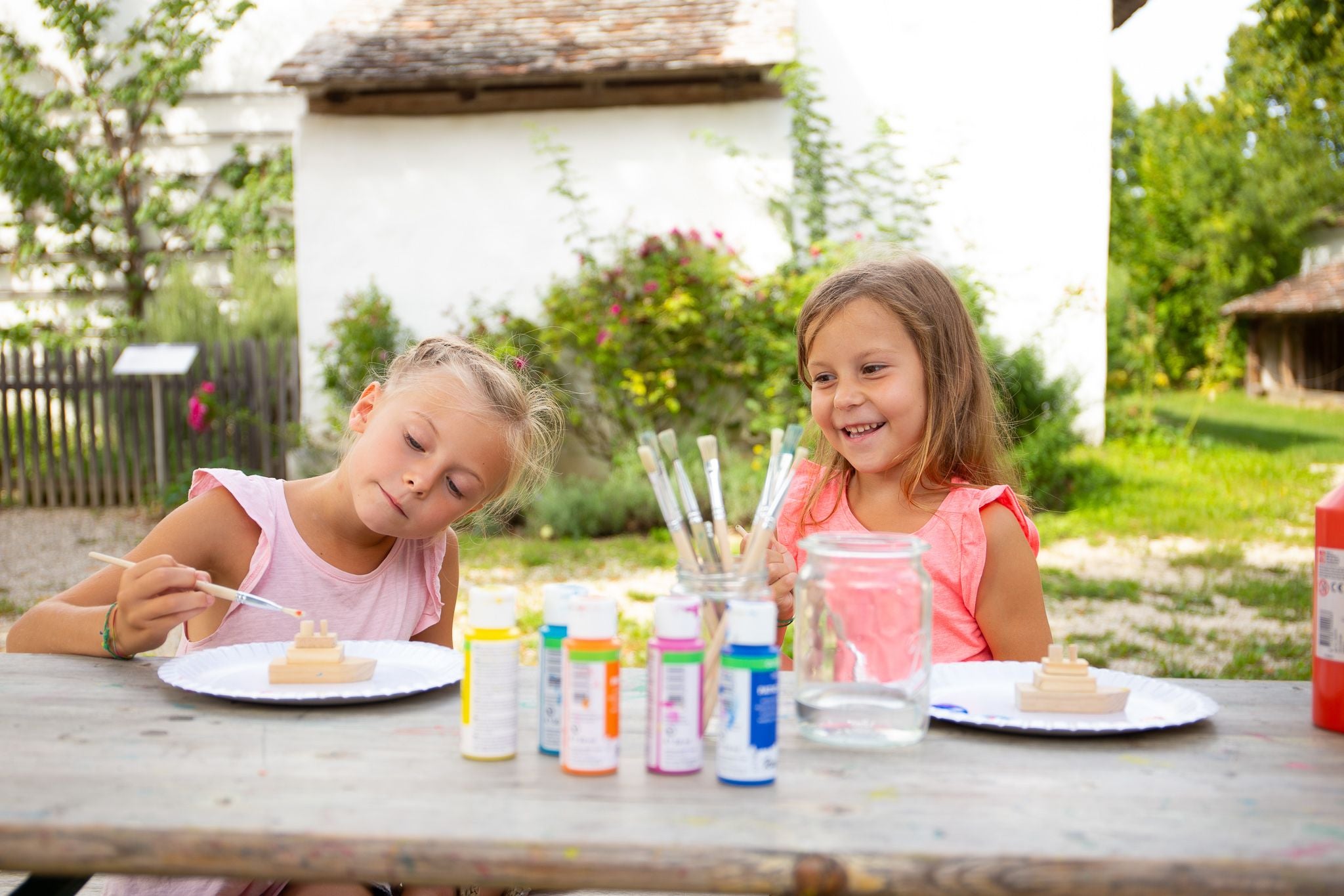 Zwei Kinder bemalen Holzteile im Freien an einem Tisch mit Farben und Pinseln.