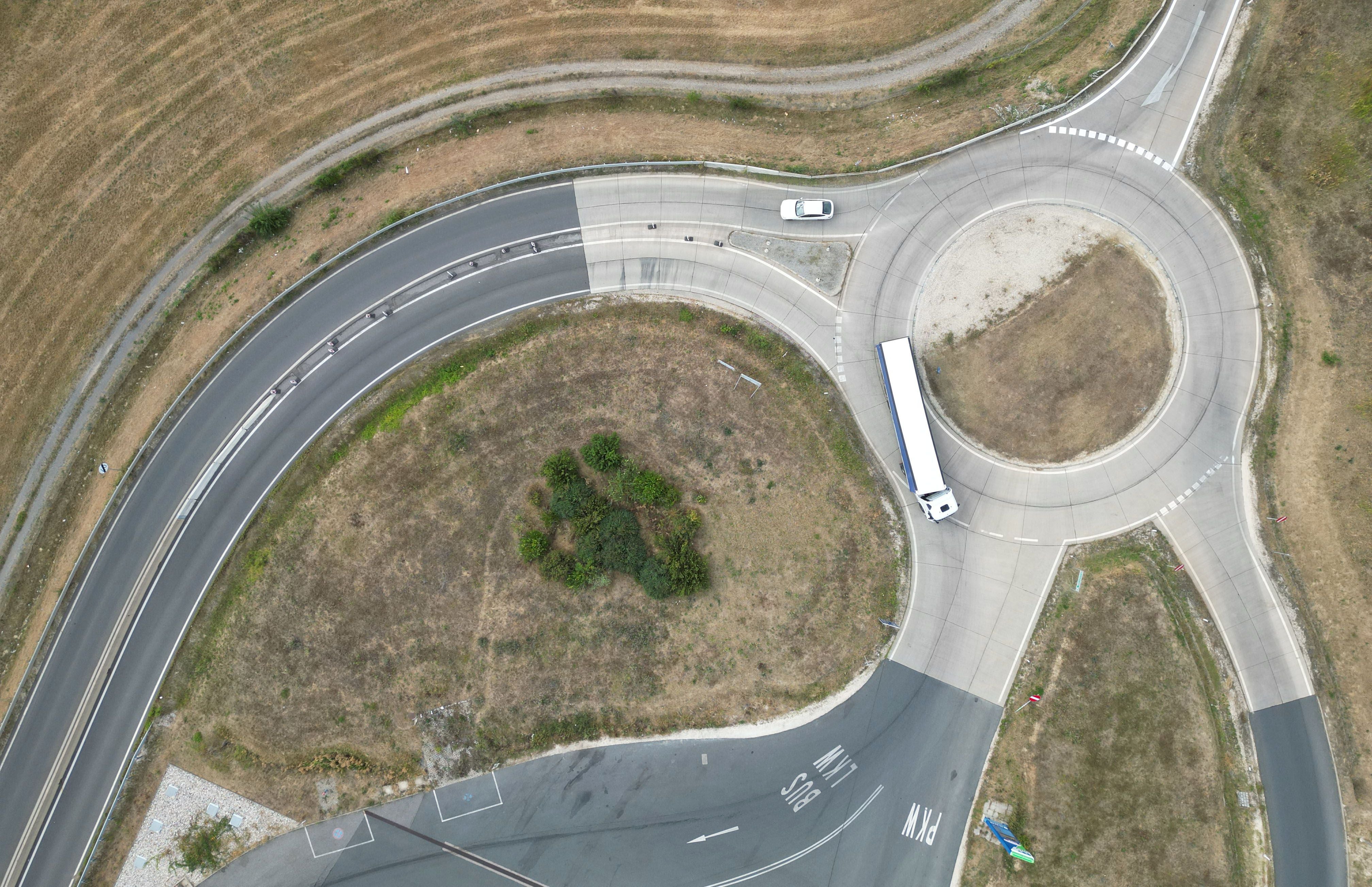 Luftaufnahme eines Kreisverkehrs mit LKW und Auto, umgeben von versiegelter Fläche und Wiese.