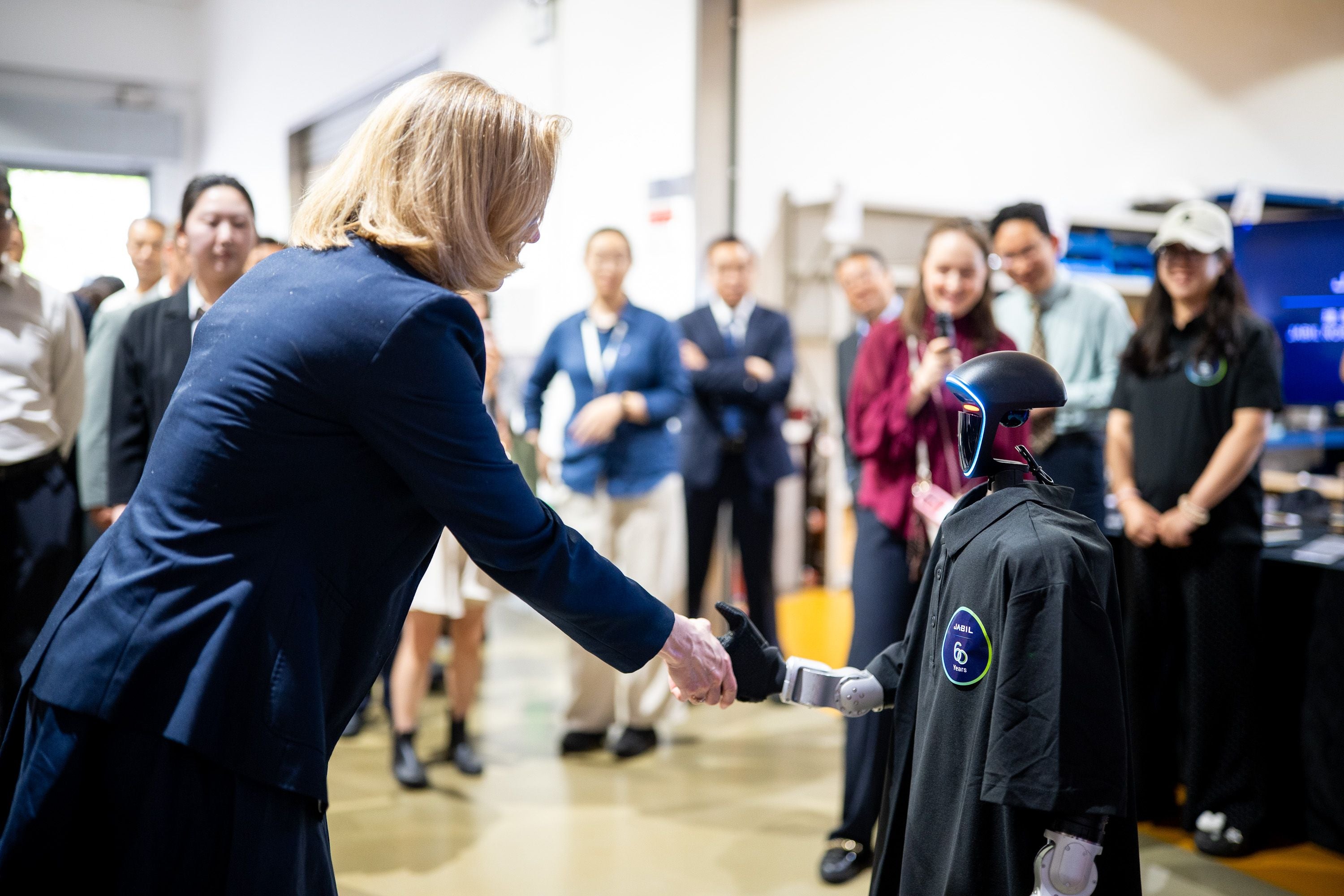 Eine Frau in Anzug schüttelt einem humanoiden Roboter vor einer Gruppe Menschen die Hand.