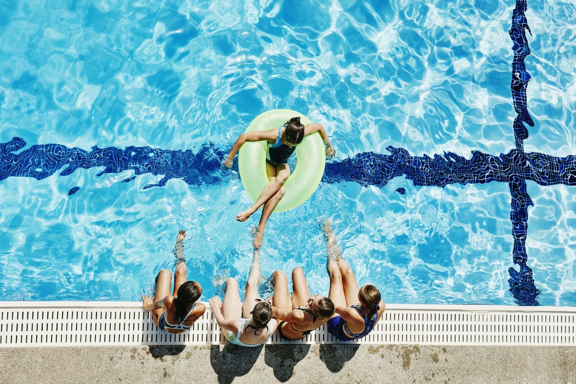 Vier Kinder sitzen am Beckenrand, eines schwimmt im Pool auf einem grünen Schwimmreifen.