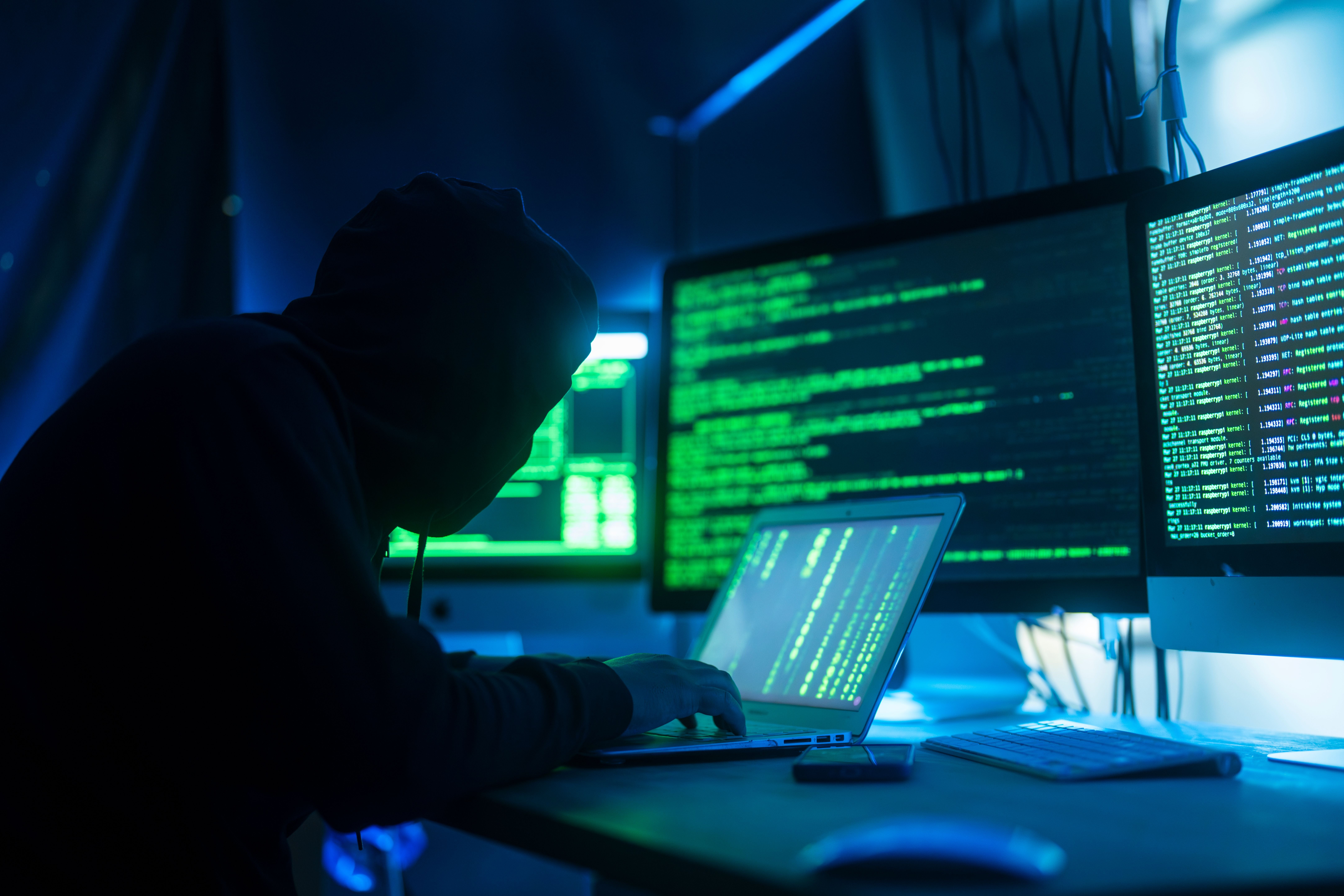 Person mit Kapuzenpulli arbeitet an Laptop mit Code auf mehreren Bildschirmen in dunklem Raum.