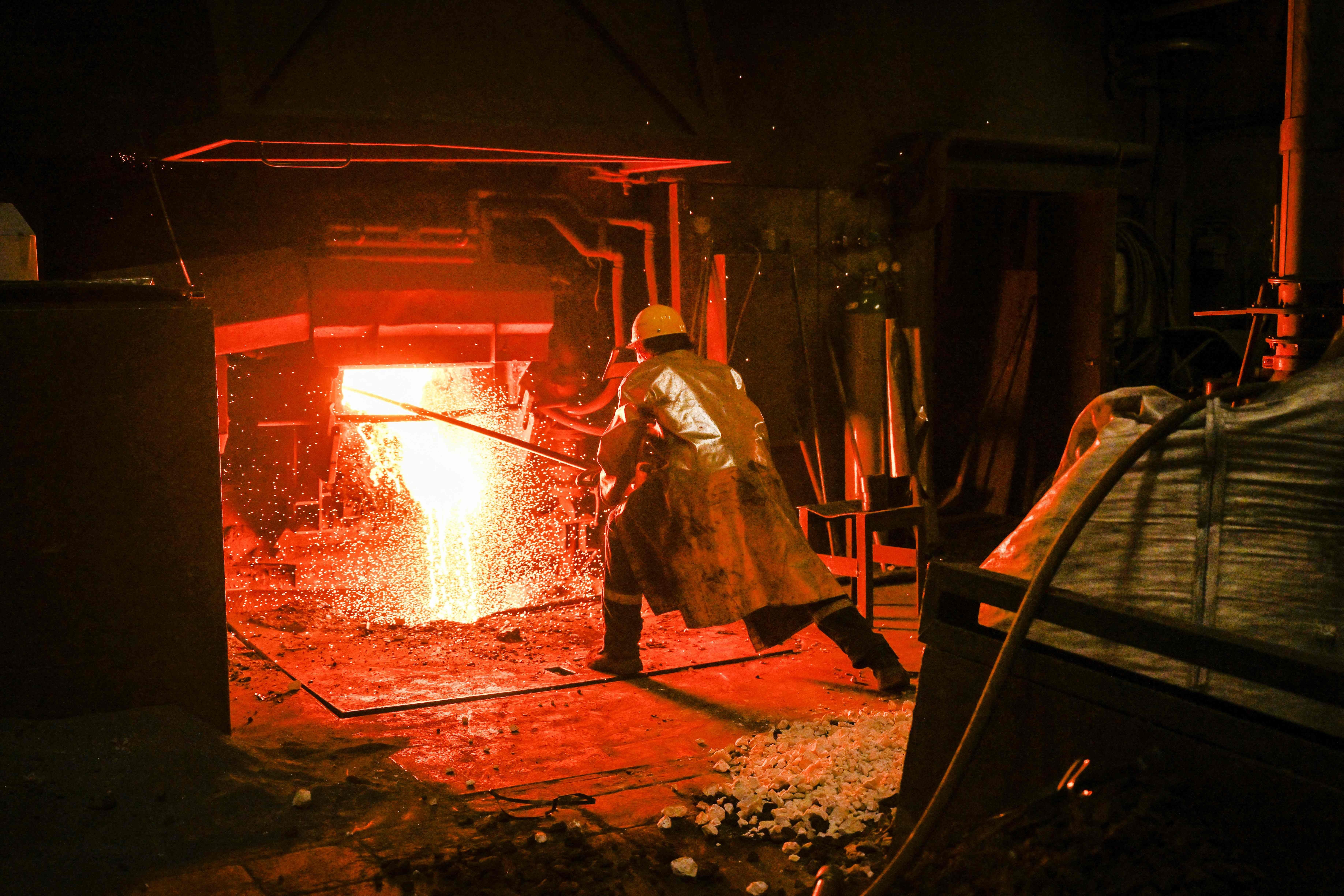 Ein Arbeiter steht in Schutzkleidung vor einem glühenden Elektroofen mit sprühendem, geschmolzenem Metall.