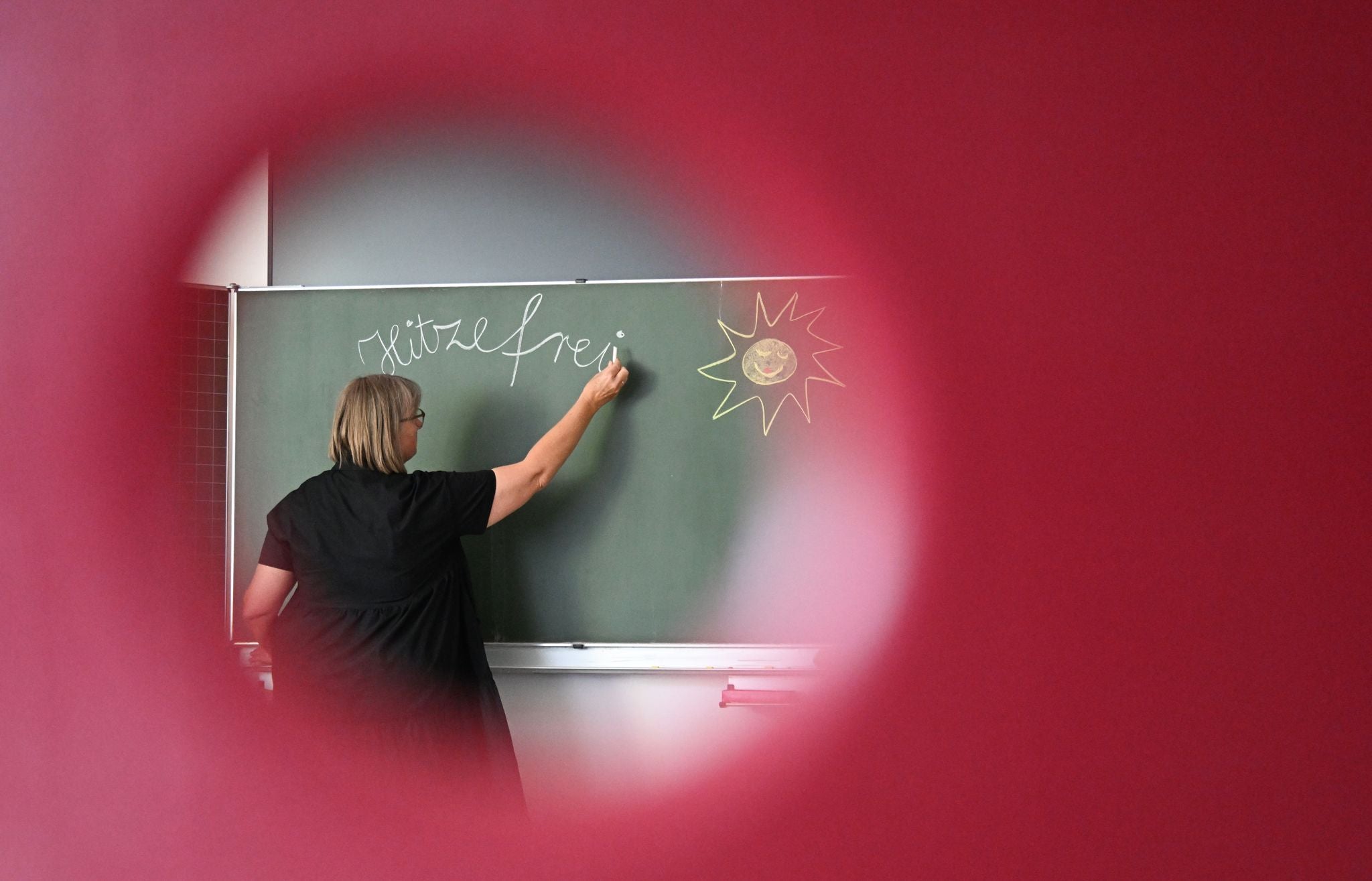 Eine Lehrerin schreibt "Hitzefrei" an eine Tafel, durch ein Loch in einem Stuhl fotografiert.