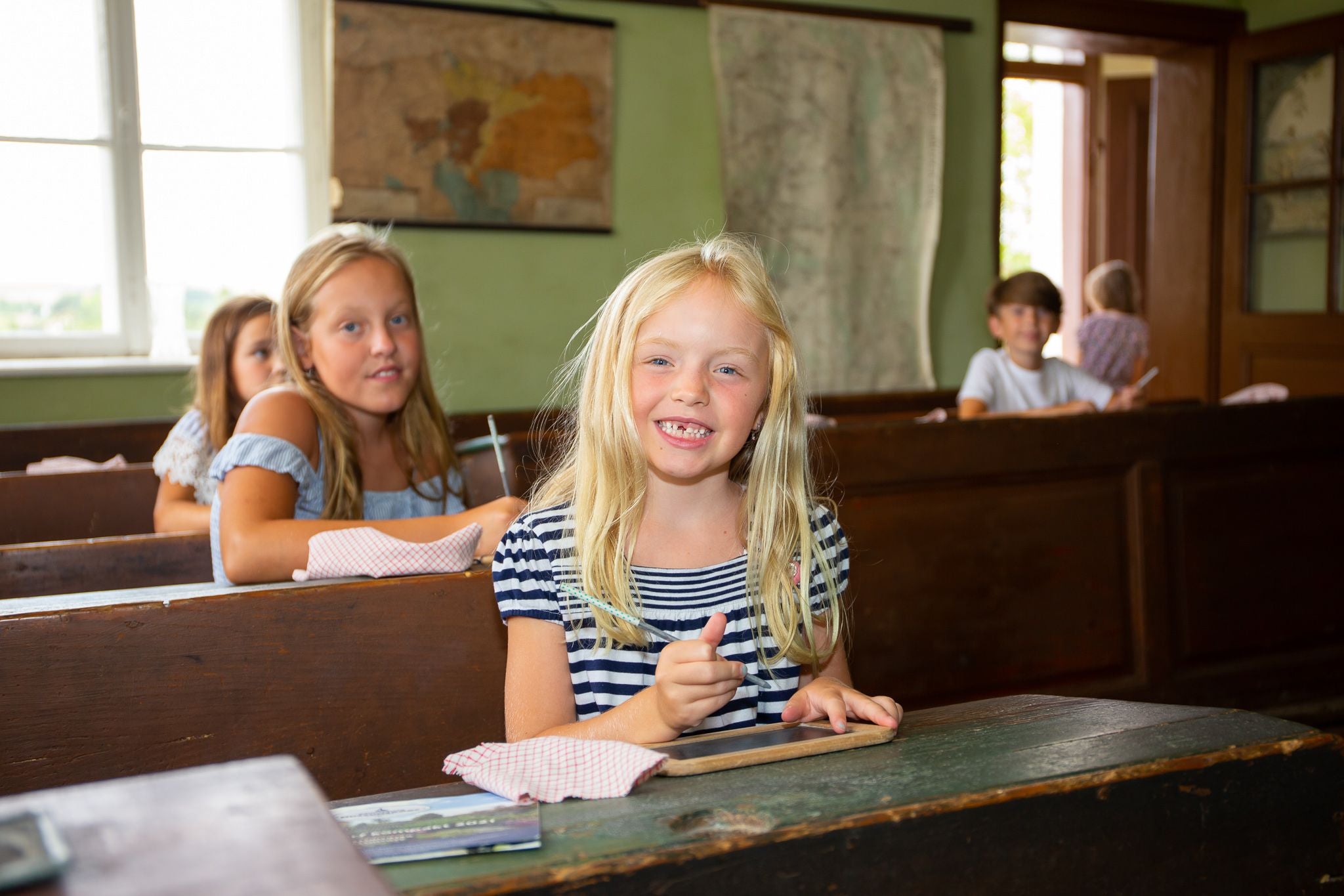 Kinder sitzen im Klassenzimmer eines Museumsdorfs und arbeiten an Schiefertafeln.