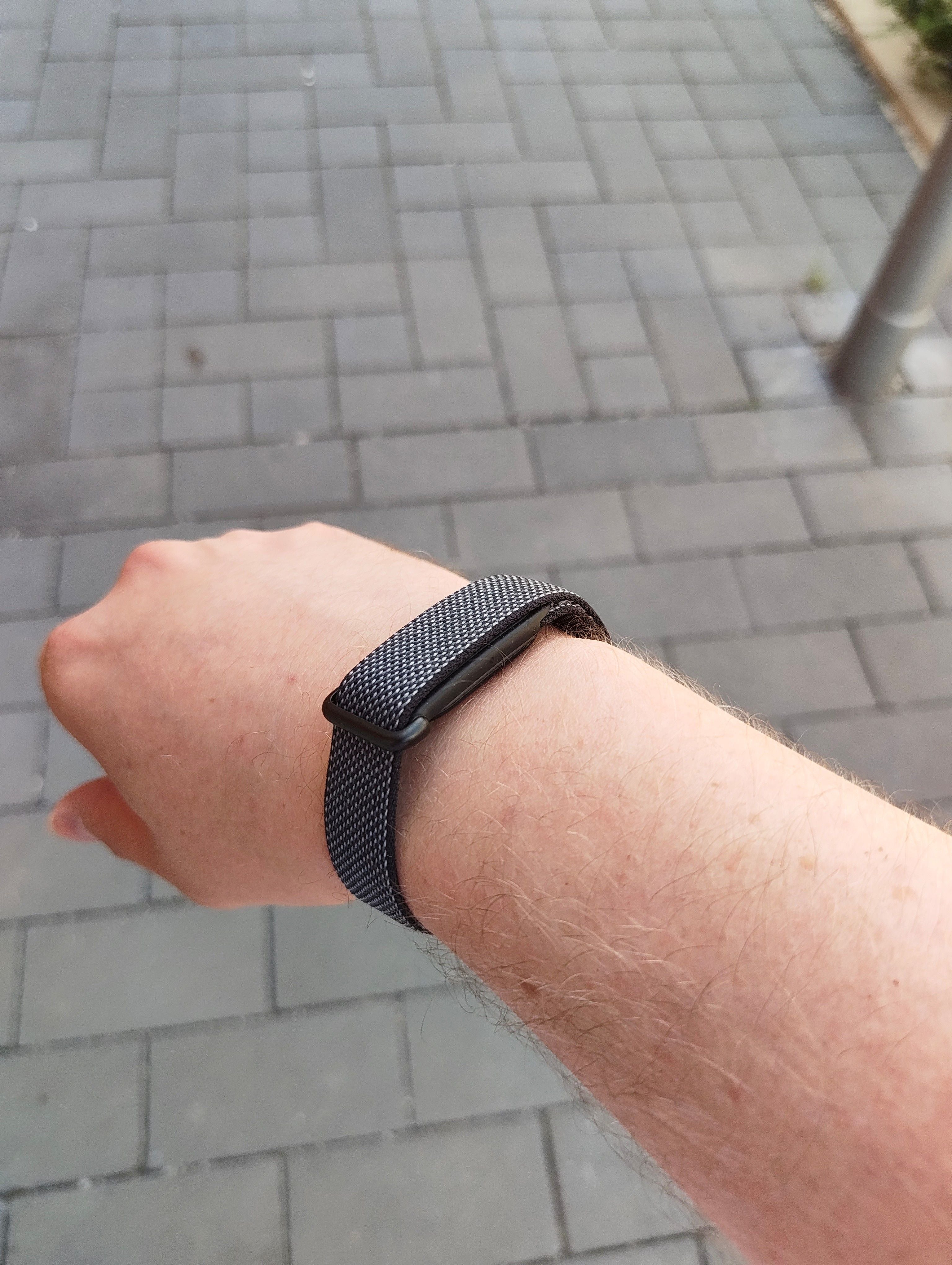 Ein Arm mit einem schwarzen Google Fitbit Air Fitnessarmband auf grauem Pflaster.