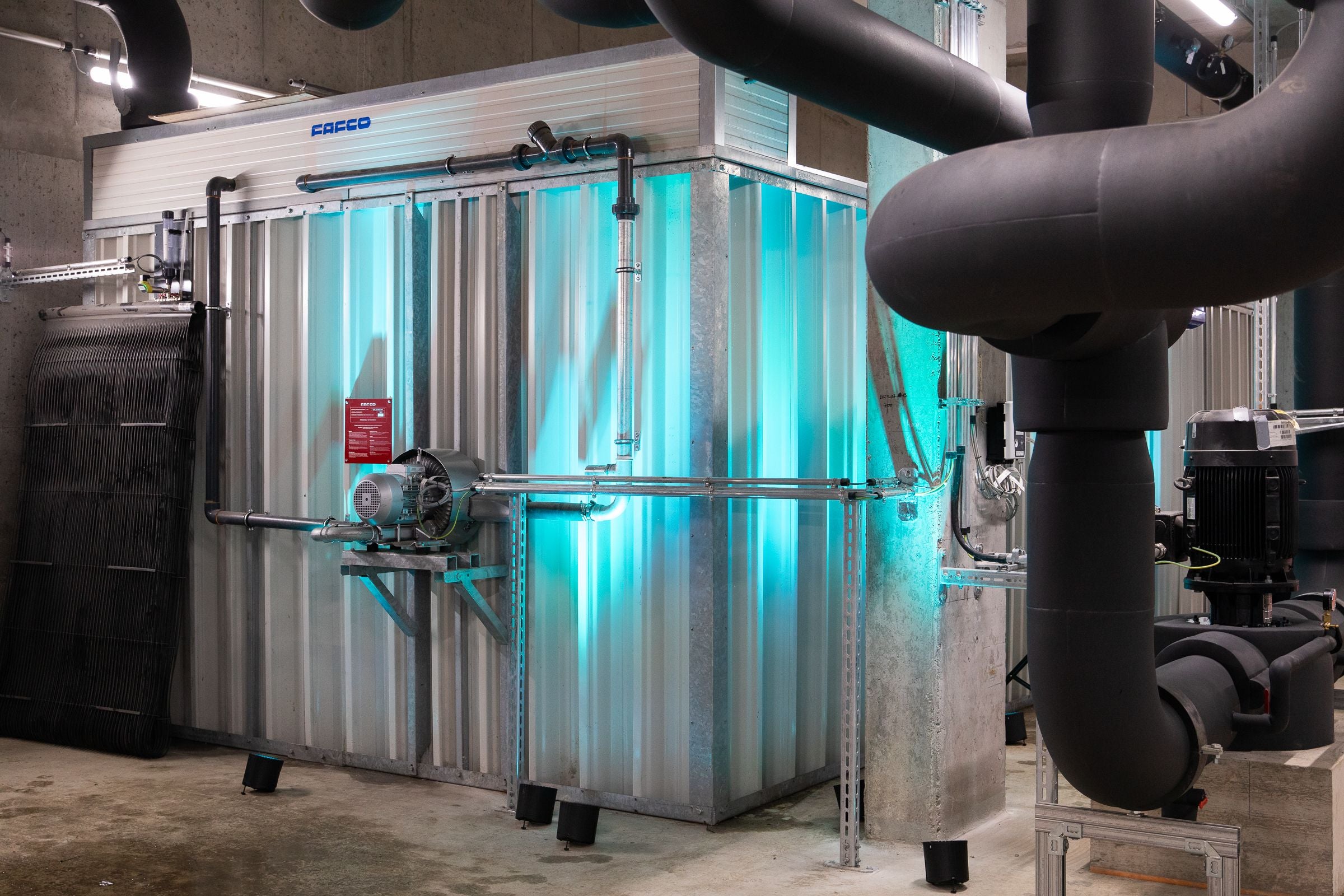 Technikraum mit Industriemaschinen, Rohren und metallverkleidetem Aggregat, blau beleuchtet.