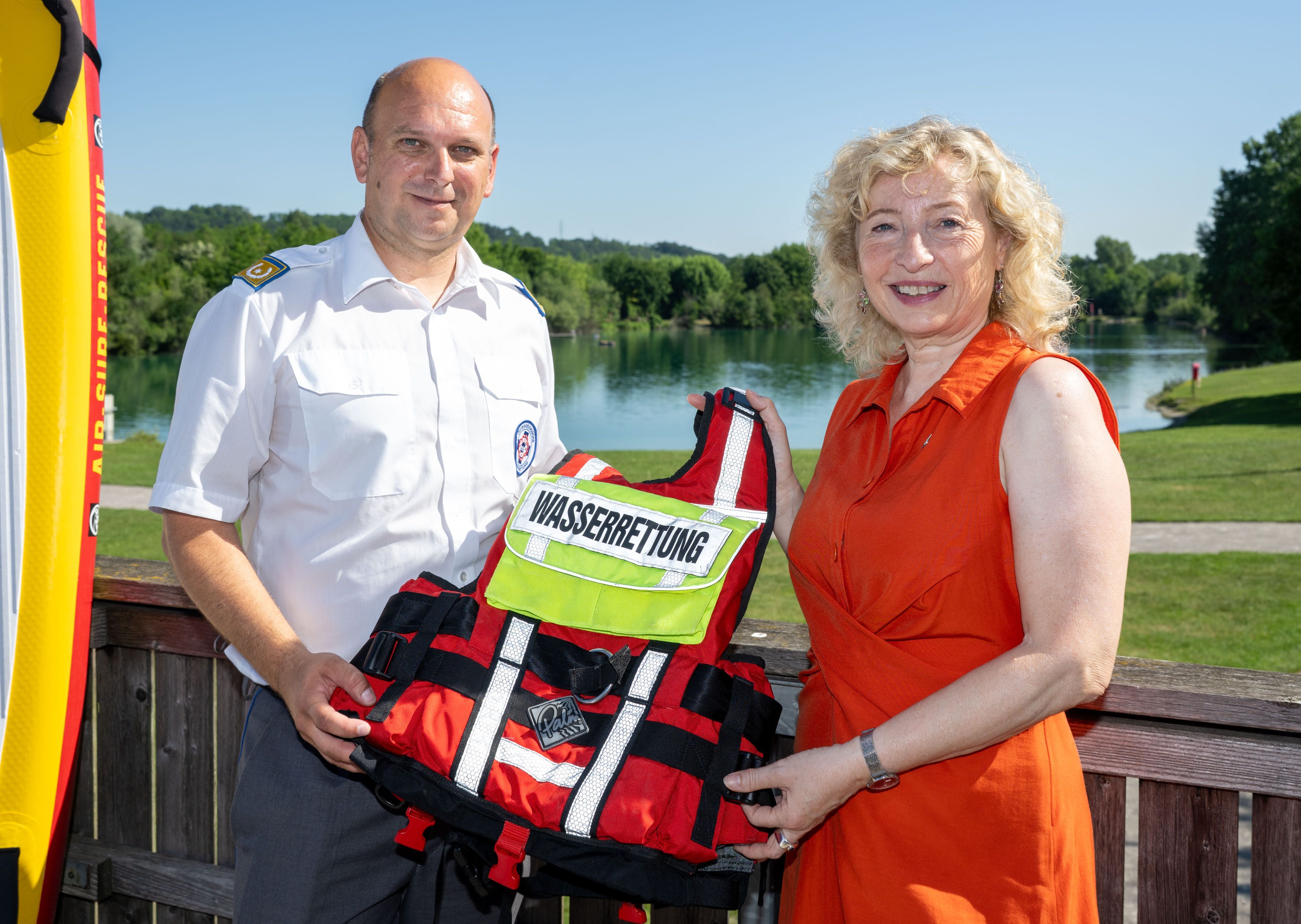 Zwei Personen halten eine Rettungsweste mit der Aufschrift „Wasserrettung“ an einem See.