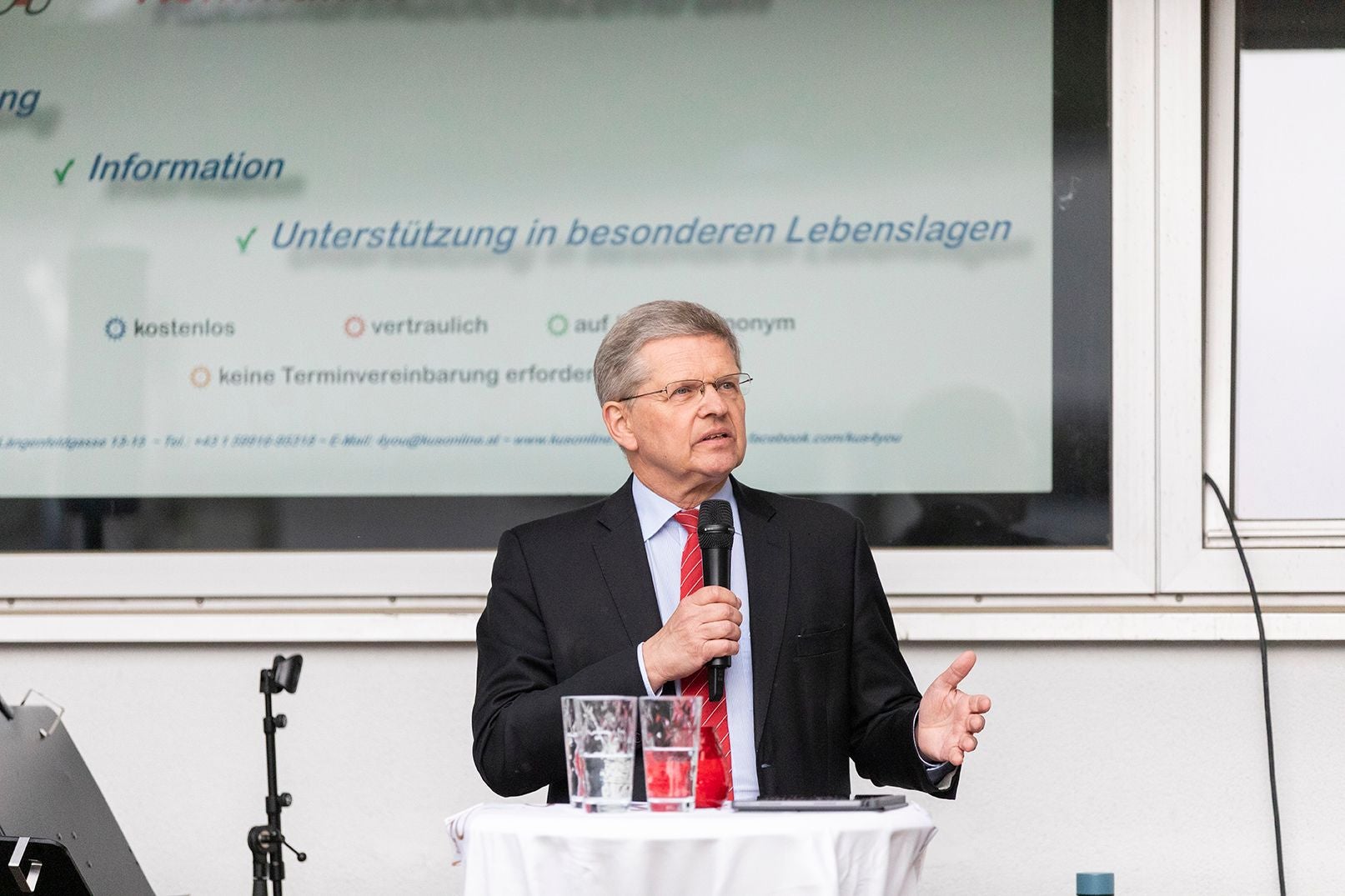 Mann im Anzug spricht mit Mikrofon vor Präsentation bei Jubiläumsfeier der VHS Meidling.