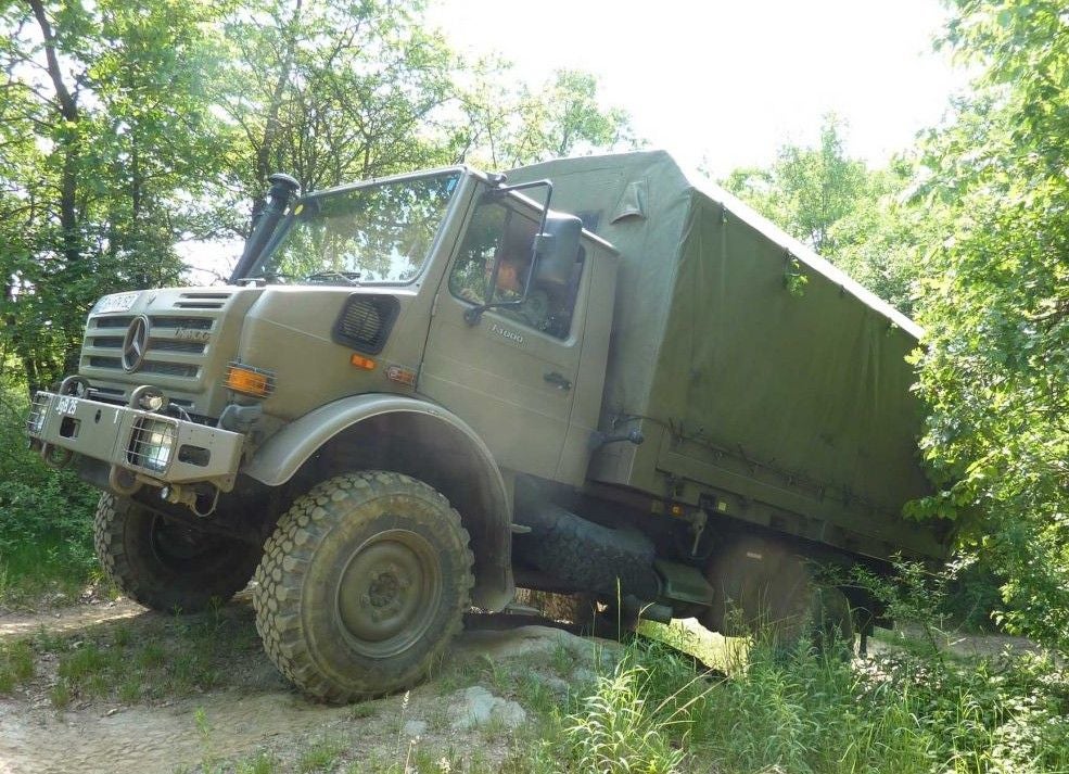 Militärischer Unimog-LKW fährt im Gelände durch bewaldetes Gebiet.