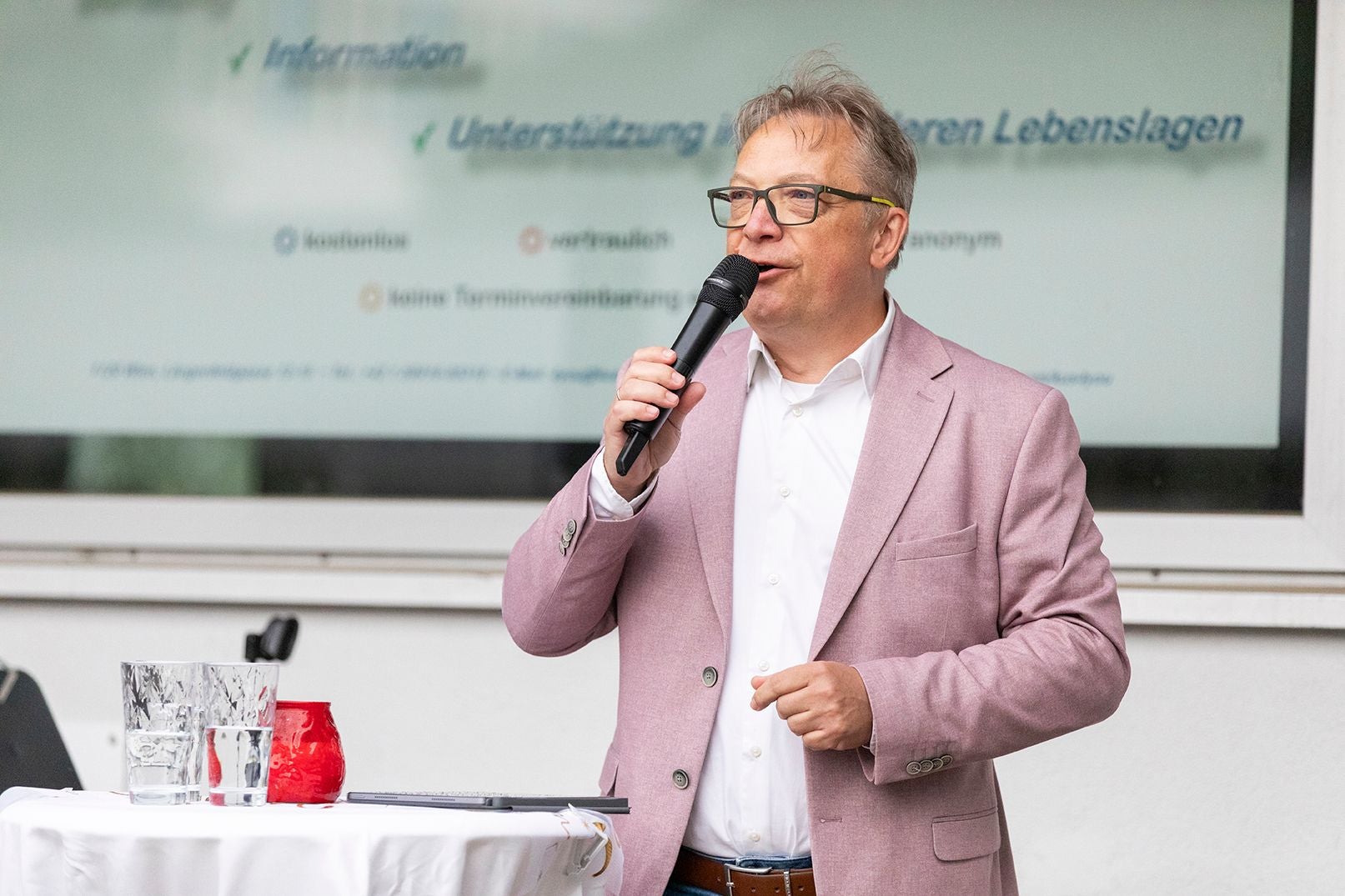 Mann im rosa Sakko spricht mit Mikrofon vor Präsentationsfolie, Tisch mit Gläsern im Vordergrund.