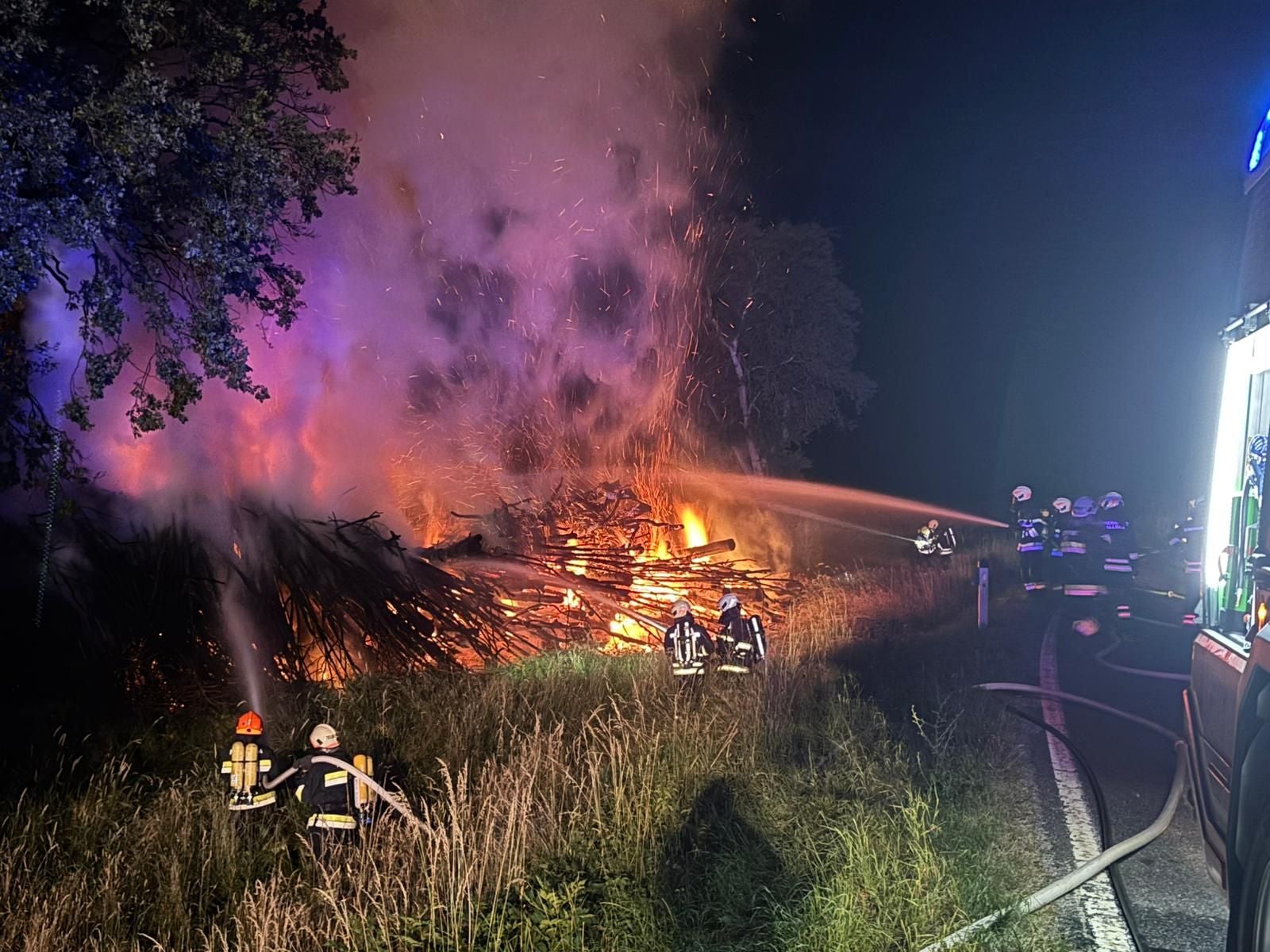 Feuerwehr löscht nachts ein großes brennendes Holzlager am Straßenrand.