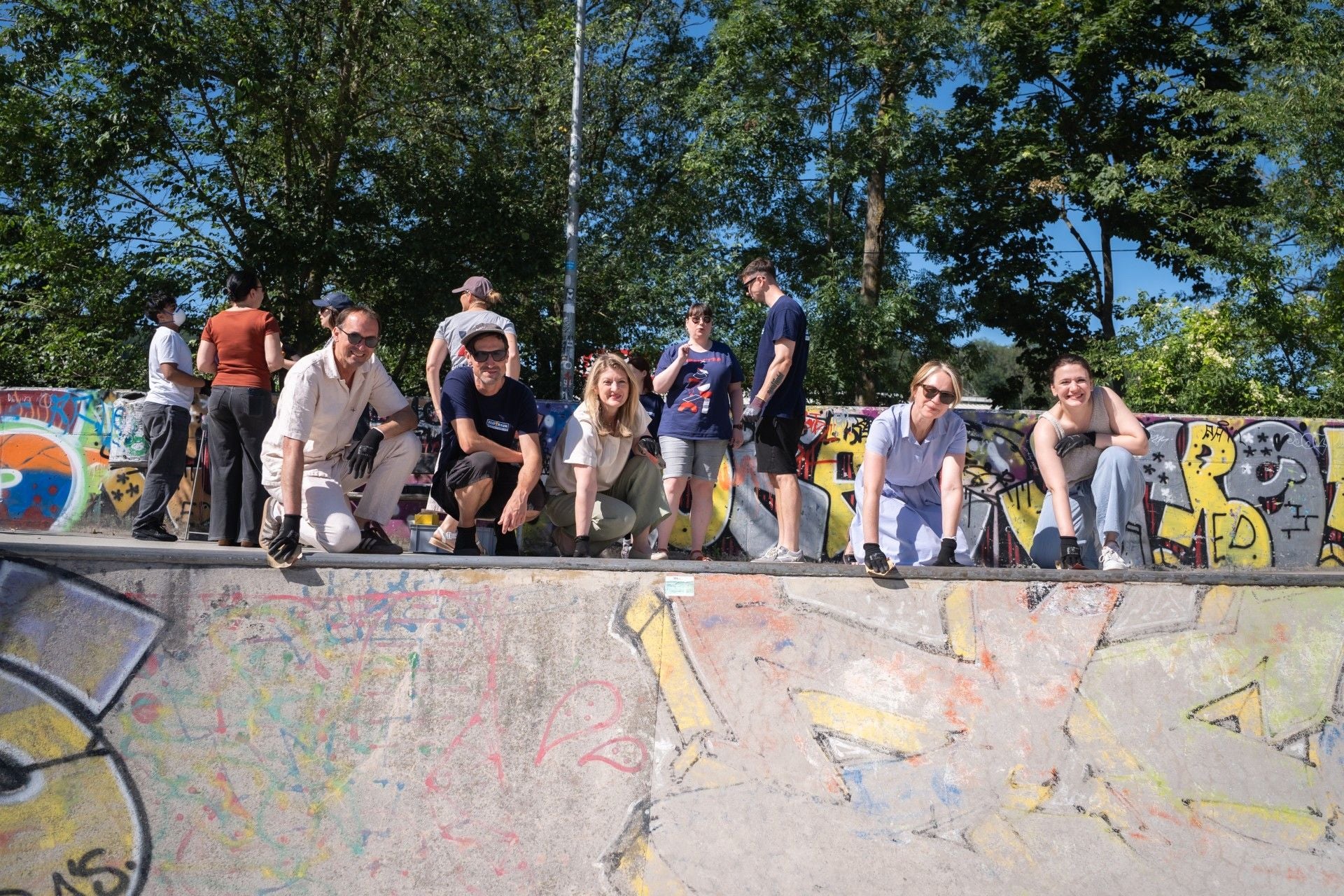 Mehrere Jugendliche renovieren einen bunt besprühten Skatepark bei sonnigem Wetter.