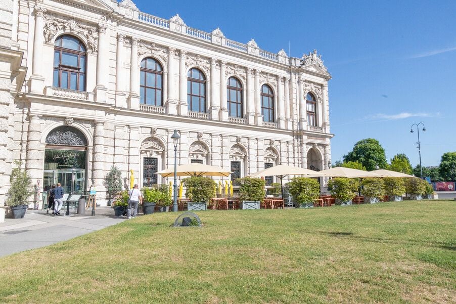 Prunkvolles Gebäude mit Terrasse, Sonnenschirmen und Tischen im Garten bei sonnigem Wetter.