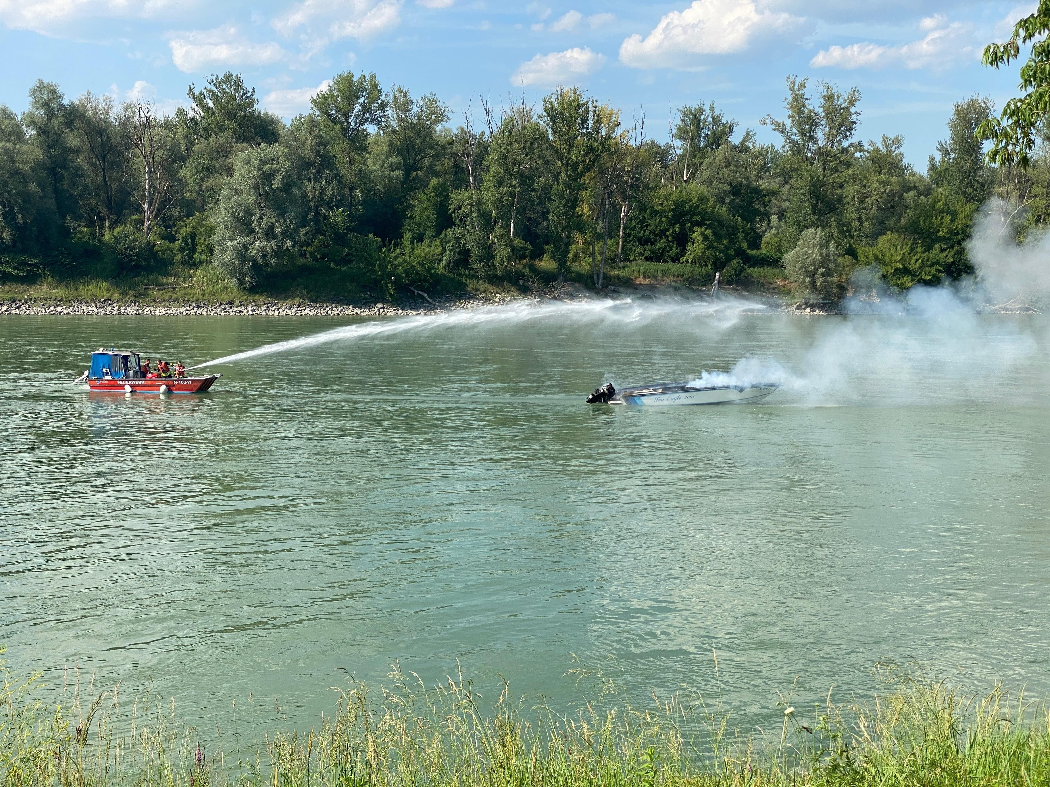 Ein Feuerwehrboot löscht mit einem Wasserstrahl ein brennendes Sportboot auf einem Fluss.