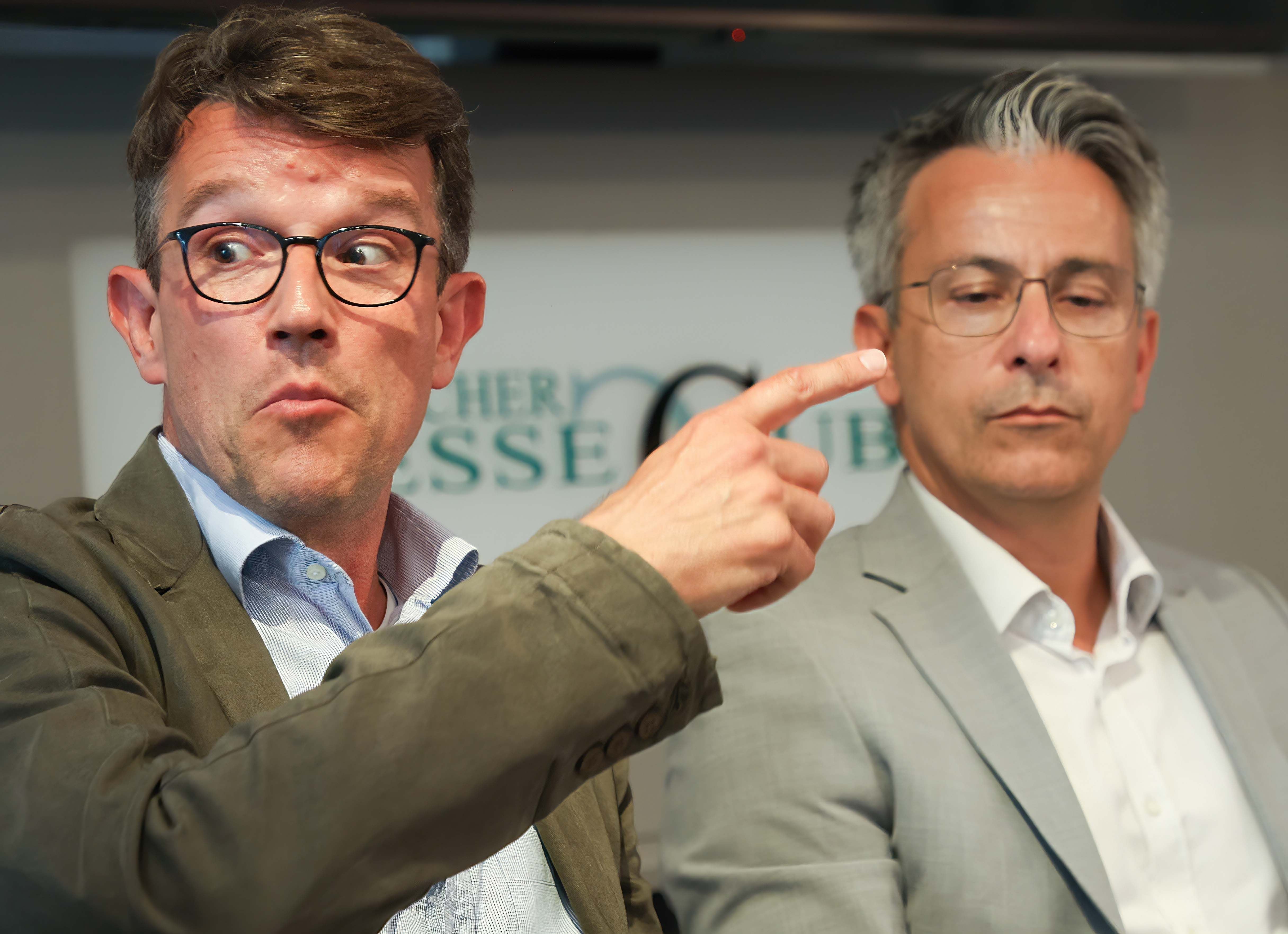 Zwei Männer im Anzug sitzen nebeneinander, einer zeigt mit dem Finger nach rechts.