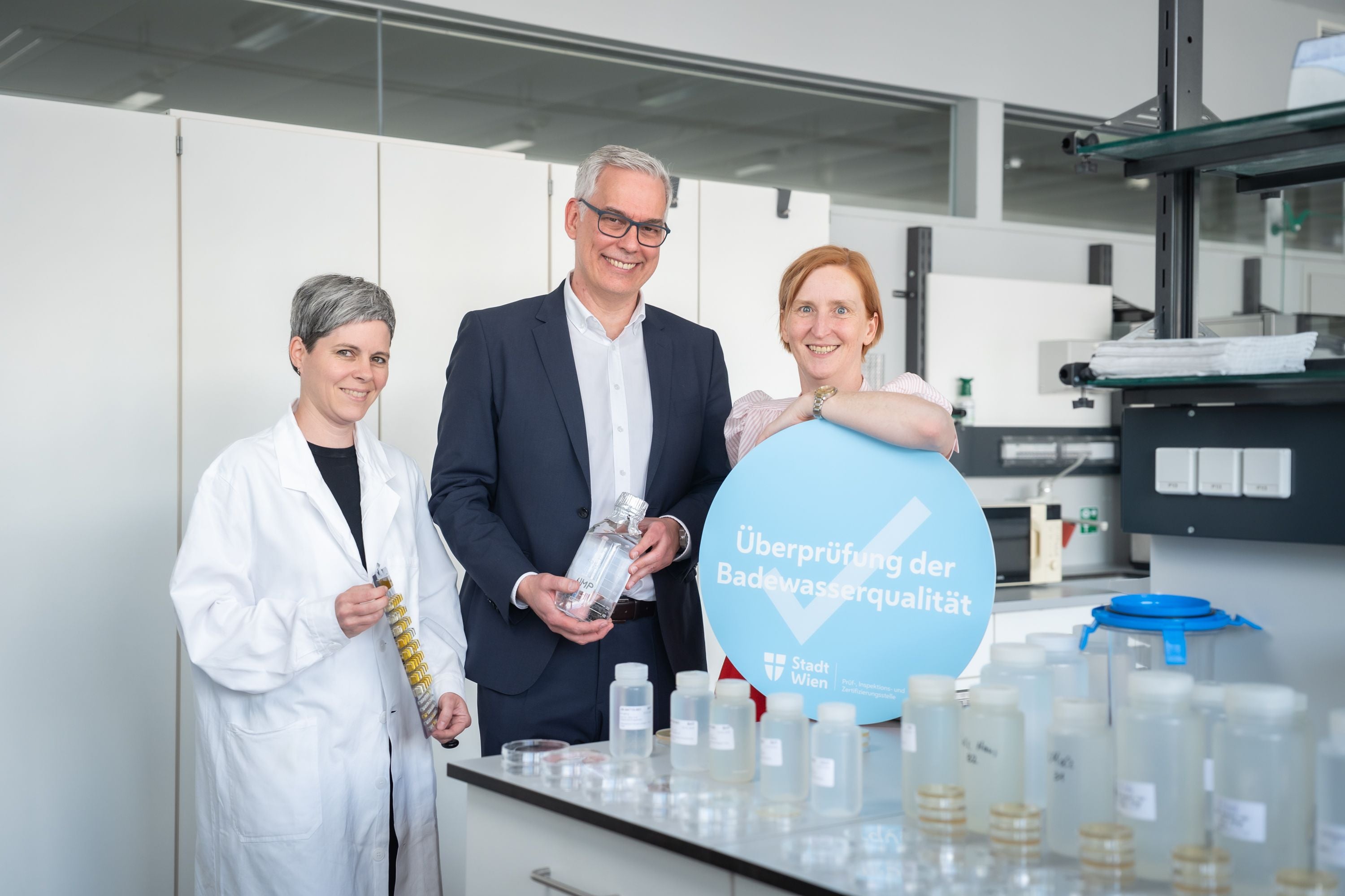 Drei Personen im Labor mit Wasserproben und einem Schild „Überprüfung der Badewasserqualität“.