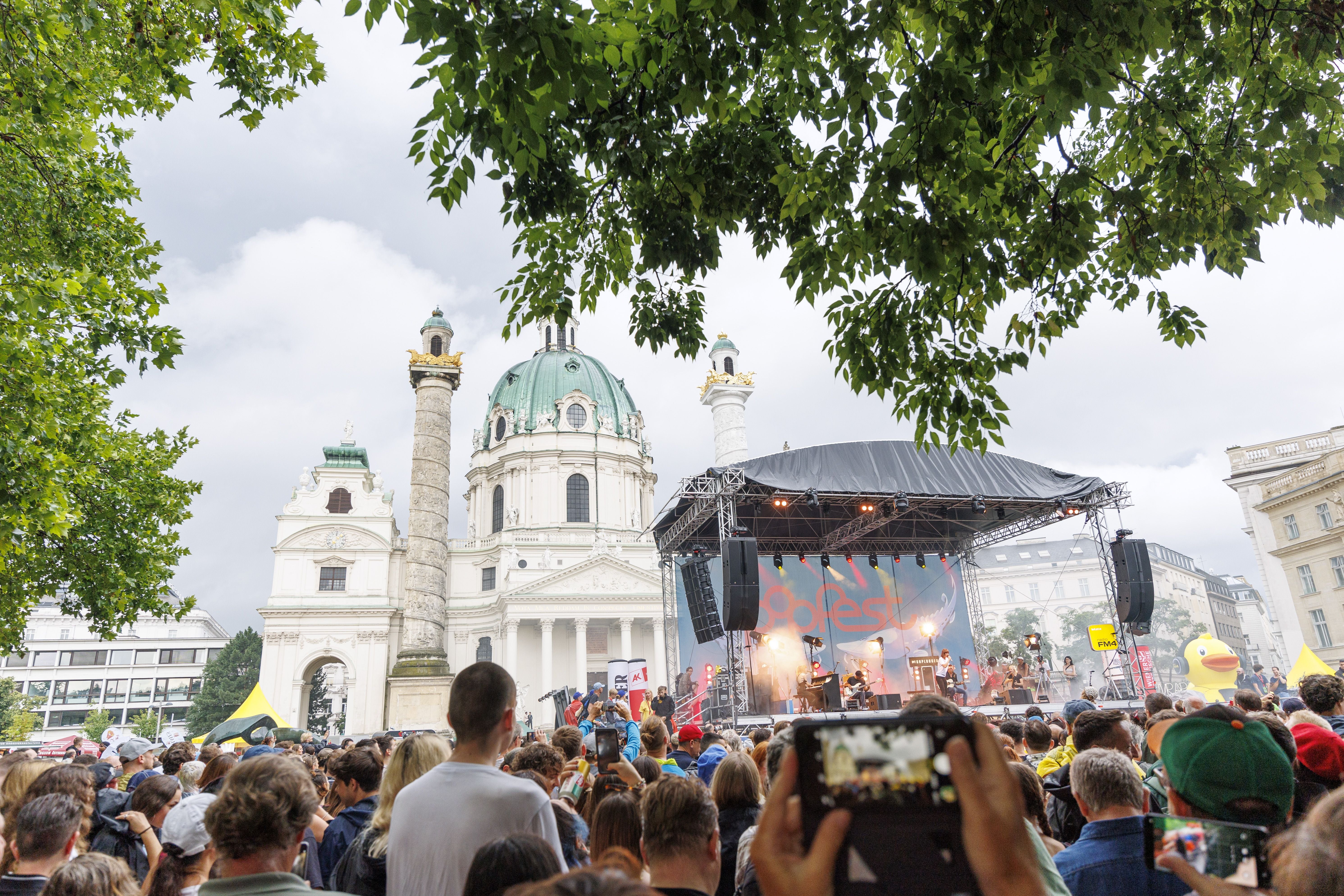 Menschenmenge vor einer Bühne beim Popfest Wien vor der Karlskirche, Sommerstimmung und Bäume.
