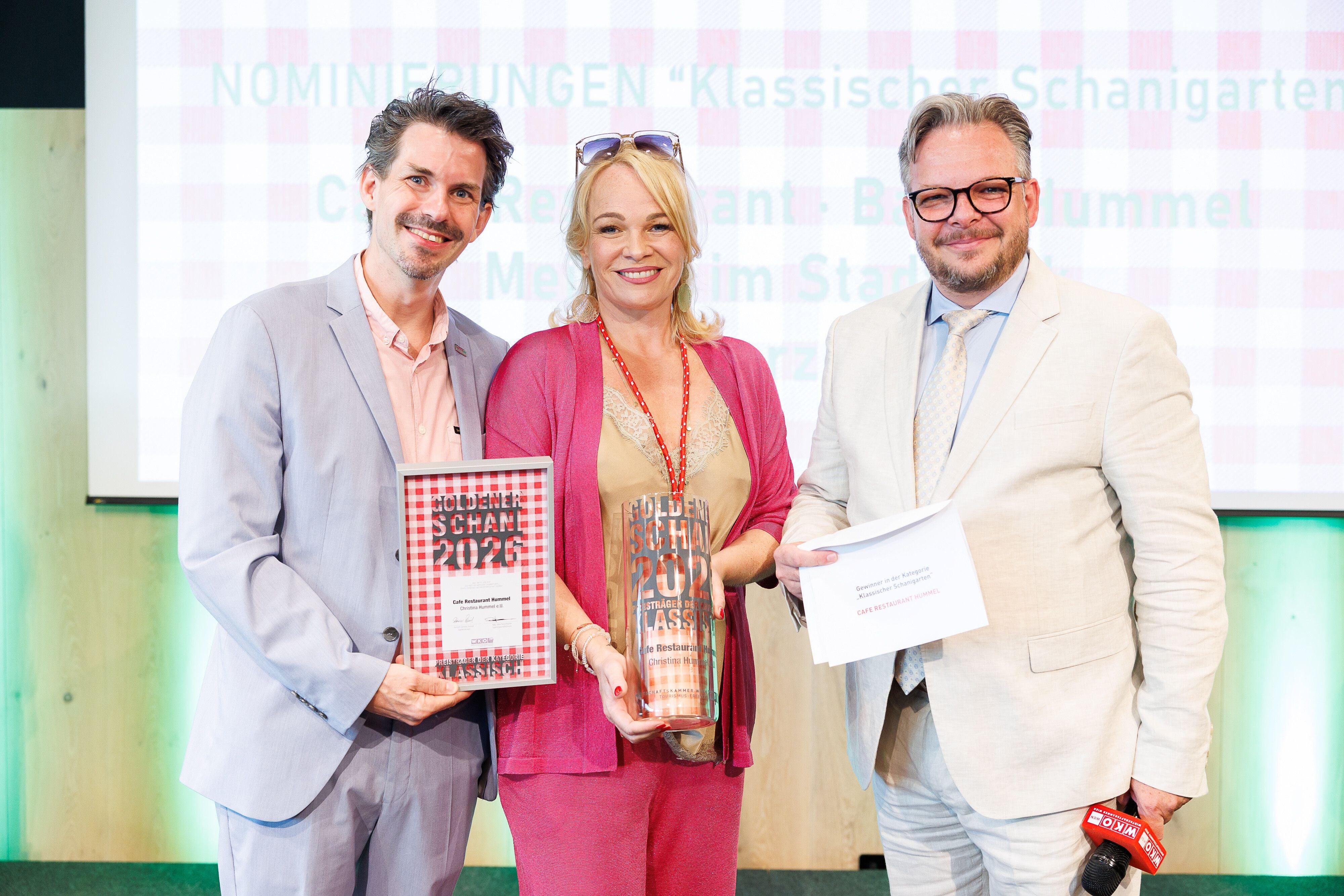 Drei Personen in festlicher Kleidung posieren mit einem Award und einer Urkunde auf einer Bühne.
