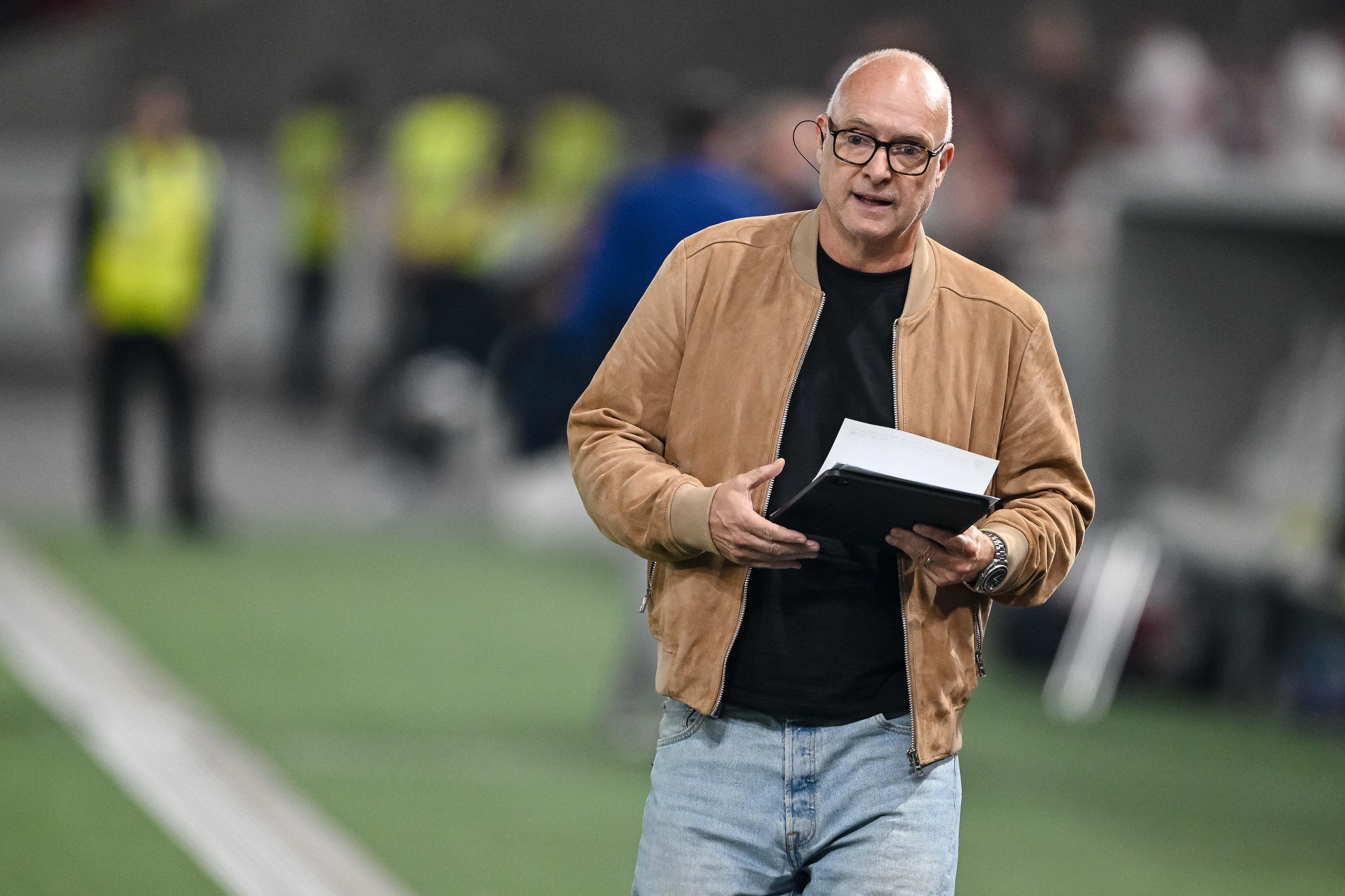 Ein Mann mit brauner Jacke hält ein Notizbuch am Spielfeldrand eines Fußballstadions.