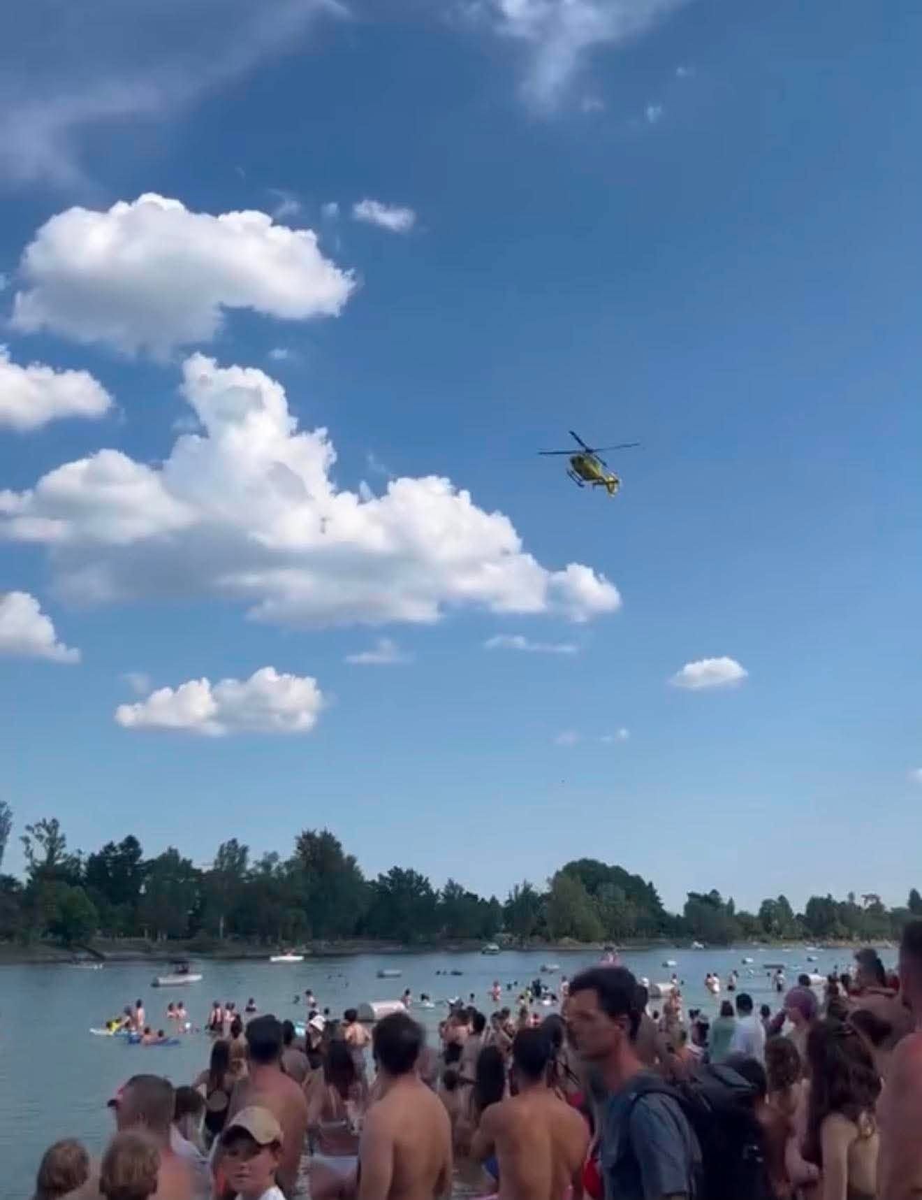 Viele Menschen stehen am Ufer, während ein Rettungshubschrauber über einem See fliegt.
