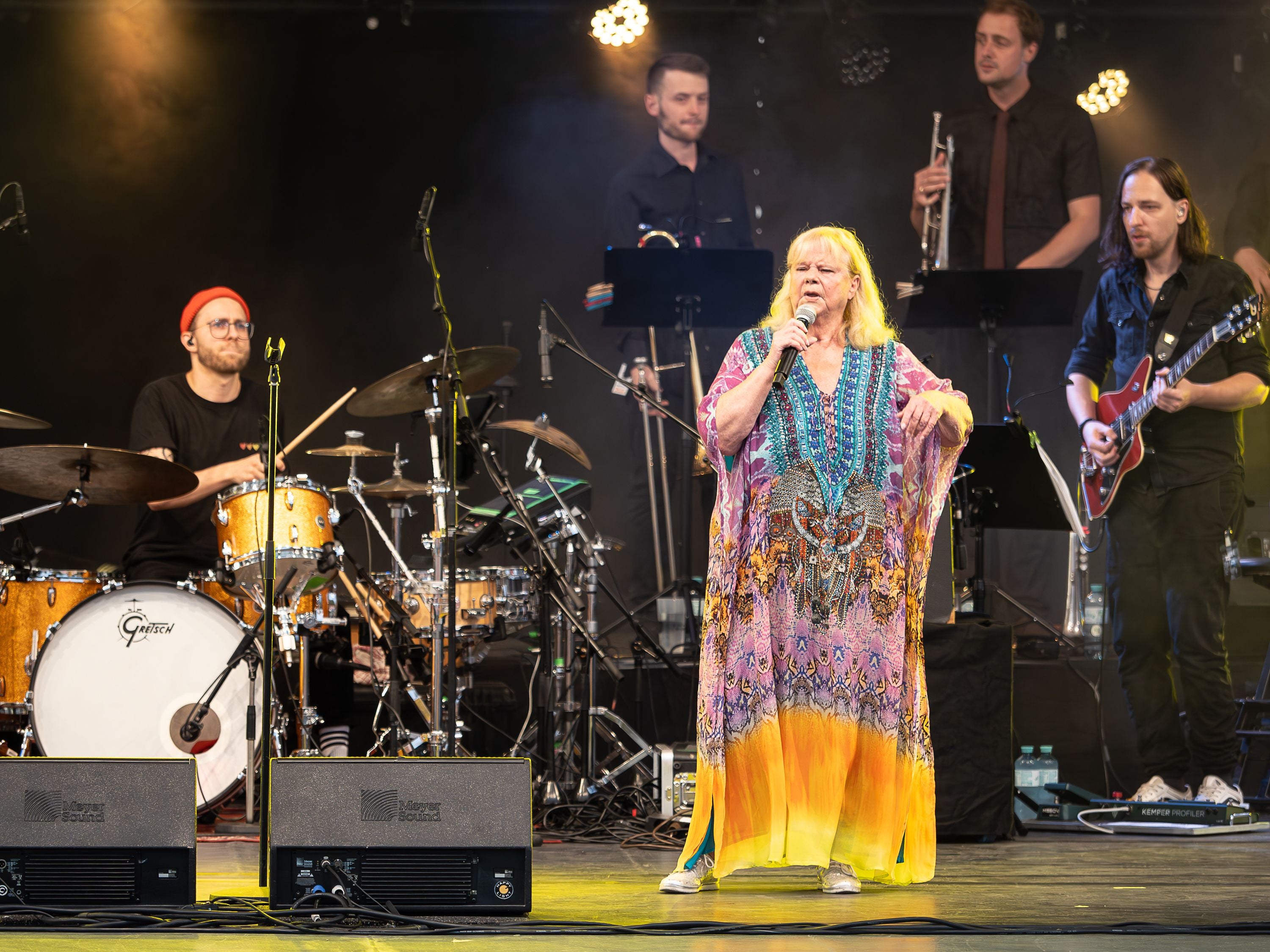 Eine Sängerin im bunten Kleid tritt mit Band auf einer Bühne mit Lichtern auf.