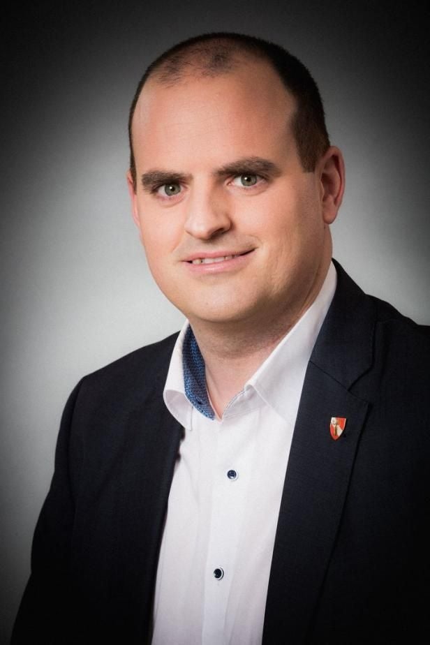 Mann im Anzug mit weißem Hemd vor neutralem Hintergrund, Gesicht unkenntlich gemacht.