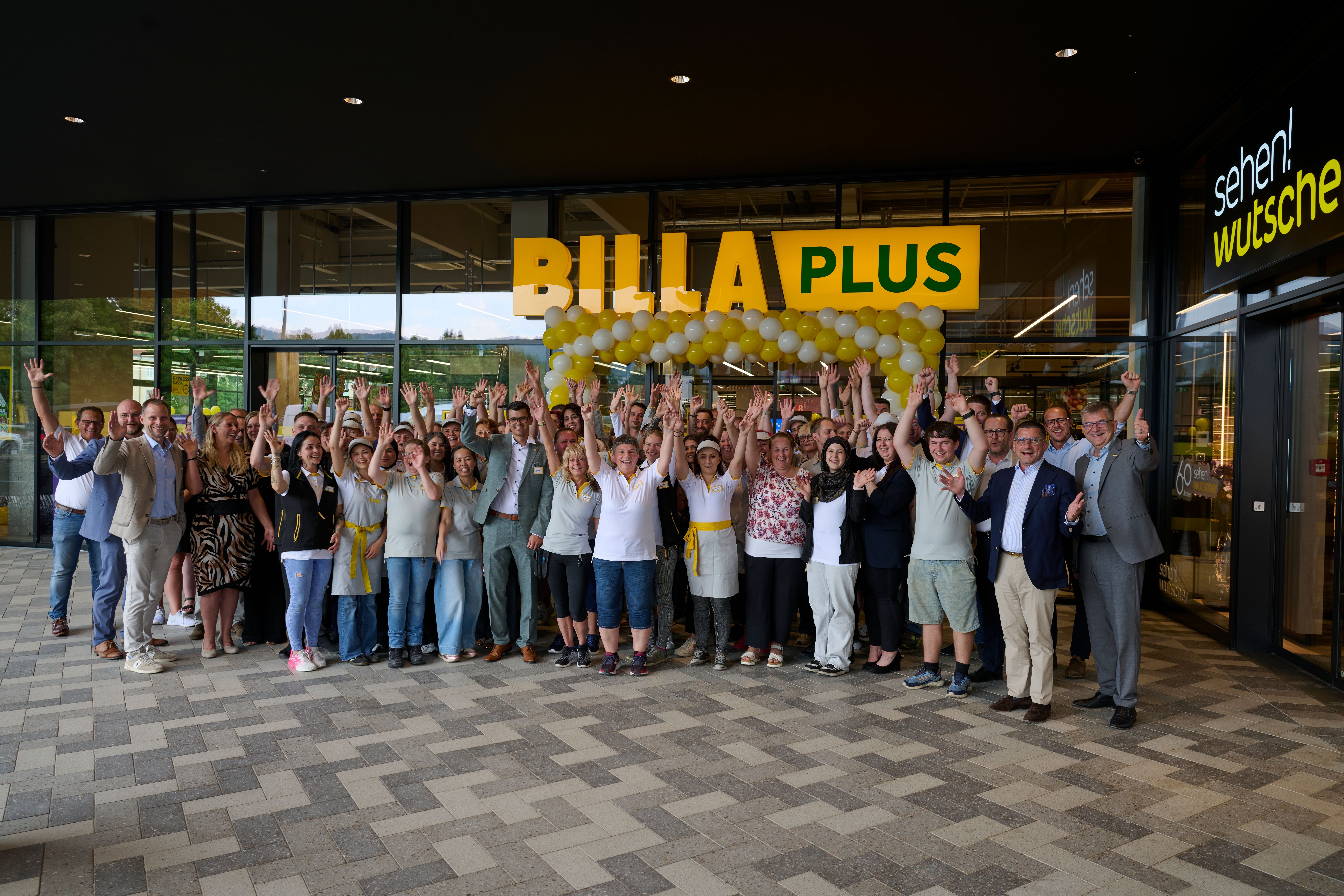 Gruppe von Menschen jubelt vor einem Billa Plus Supermarkt mit gelben und weißen Luftballons.