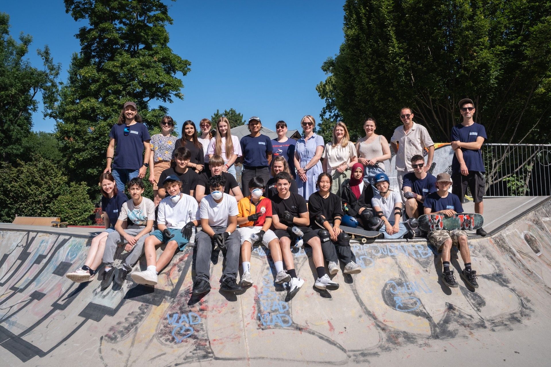 Gruppe Jugendlicher sitzt und steht im Skatepark, einige halten Skateboards, sonniges Wetter.