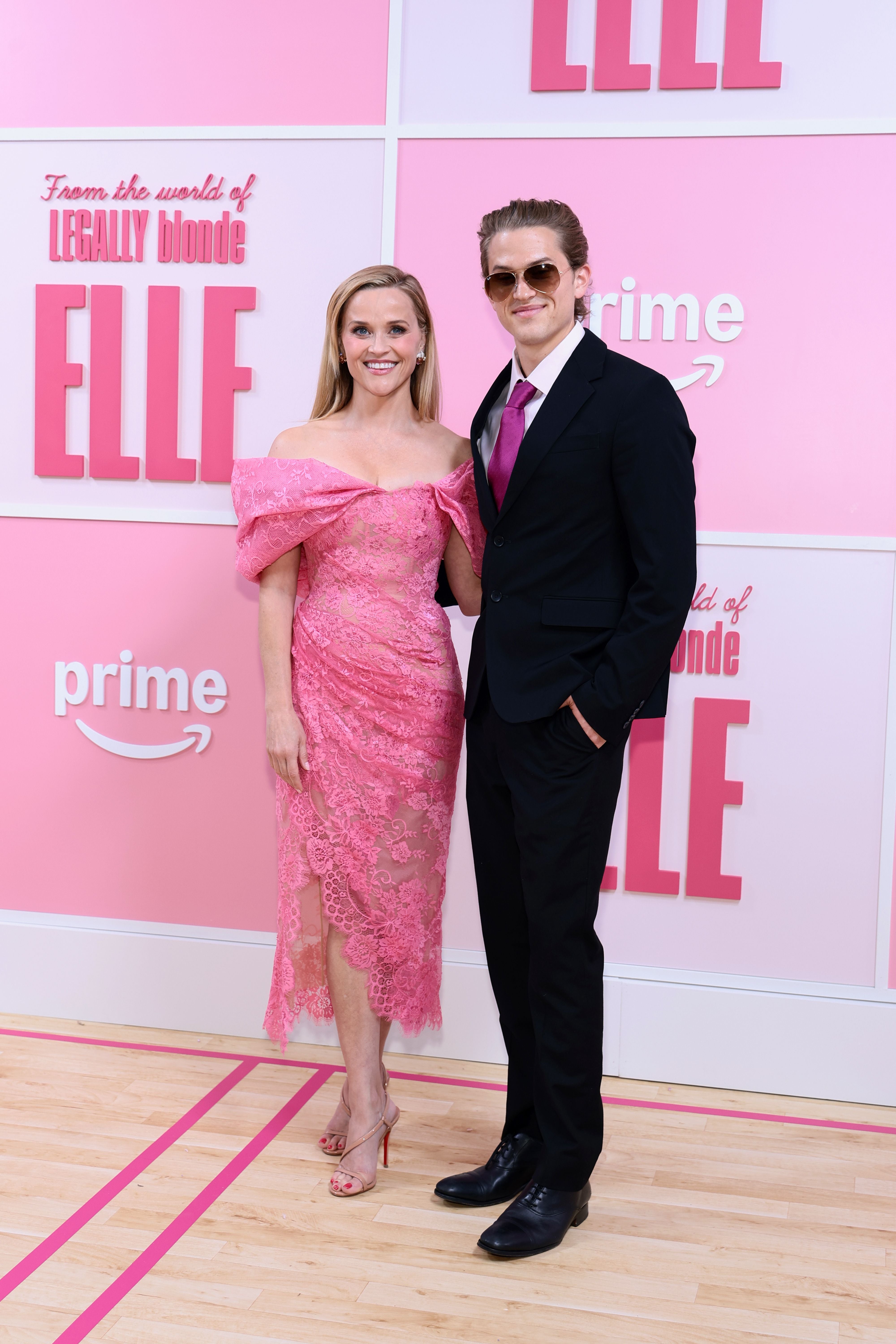 Eine Frau im pinken Kleid und ein Mann im Anzug posieren vor einer pinken „Elle“-Premierenwand.
