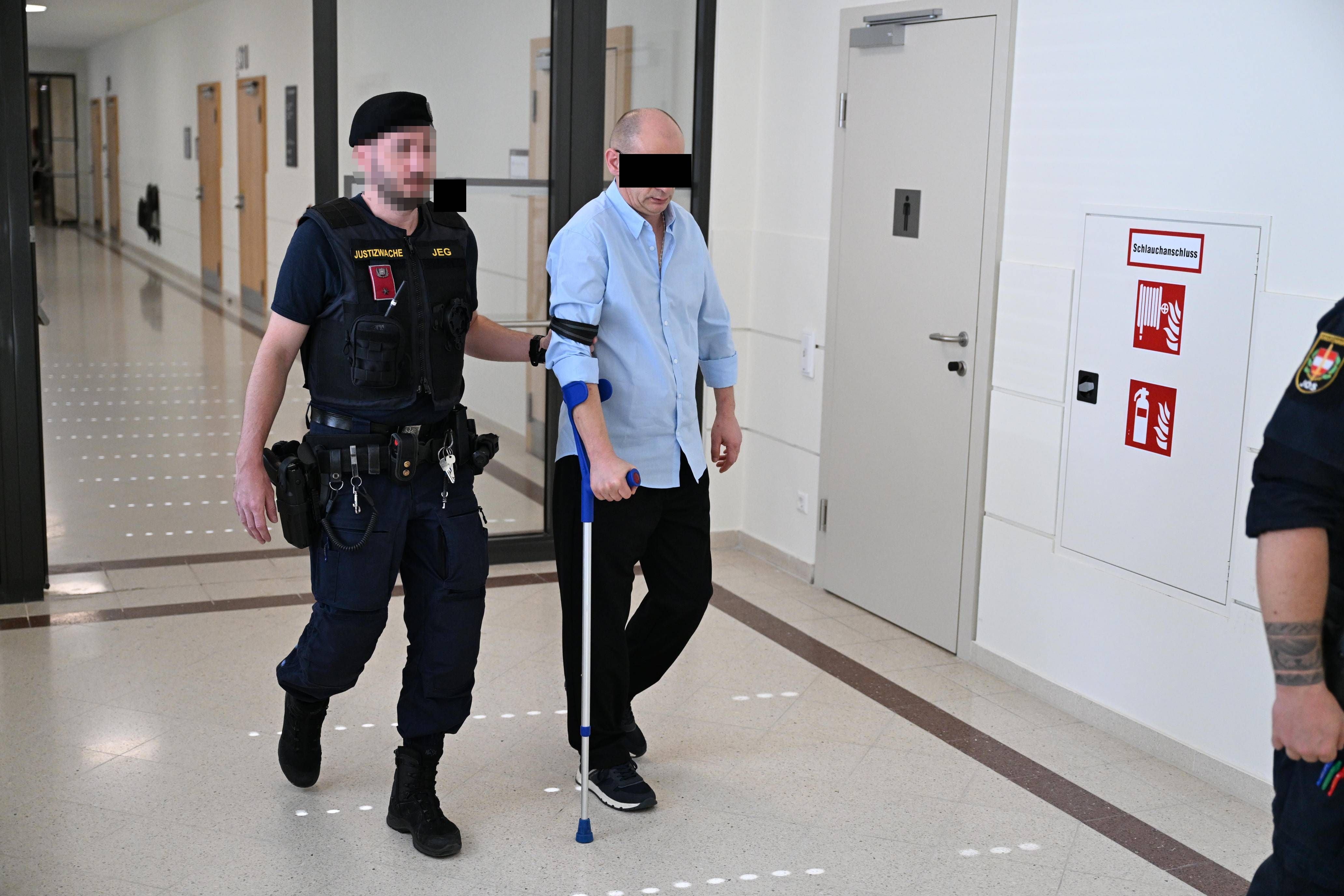 Ein Polizist führt den Angeklagten auf Krücken durch einen Gerichtsflur in Wien.