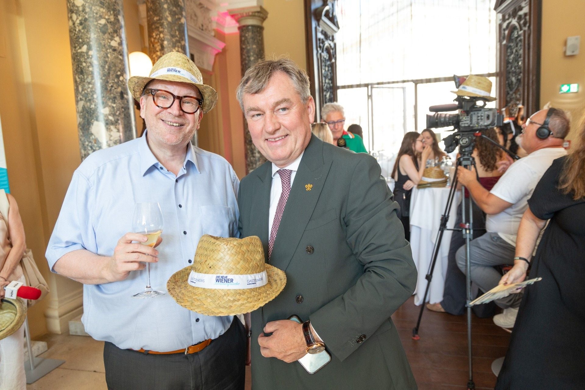 Zwei Männer mit Strohhüten posieren bei einer Sommerveranstaltung in elegantem Ambiente.