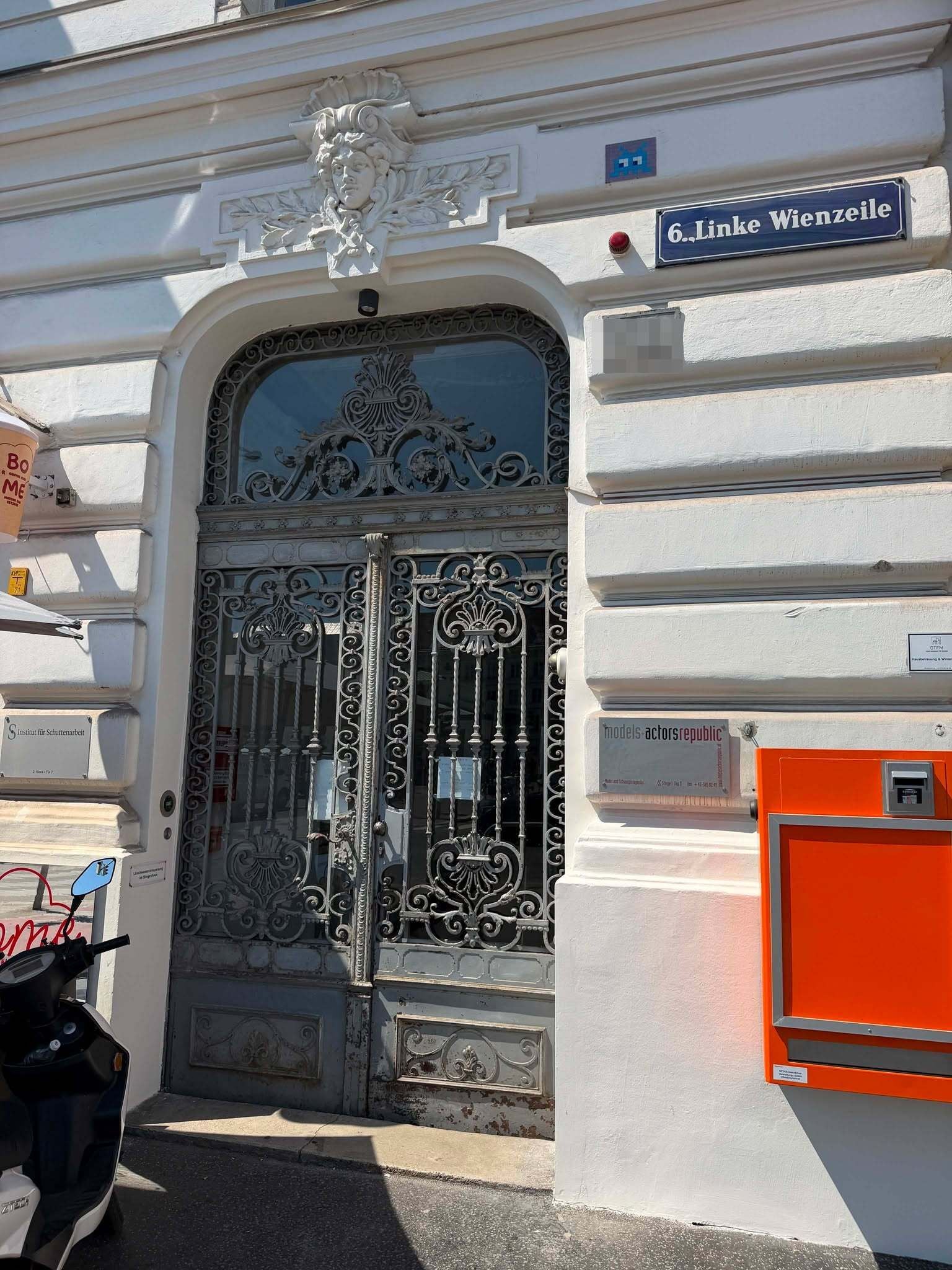 Eingang mit verzierten Eisentüren und Straßenname „6. Linke Wienzeile“ an weißer Fassade.