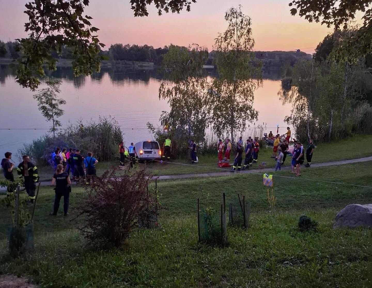 Viele Einsatzkräfte stehen an einem See, in dem ein Auto mit Anhänger halb im Wasser steckt.