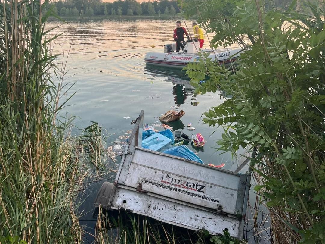 Ein Anhänger liegt halb im Wasser, umgeben von Müll, im Hintergrund ein Rettungsboot mit zwei Personen.