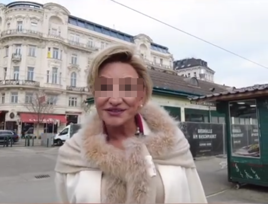 Eine Person im hellen Mantel steht auf einem Platz vor einem historischen Gebäude.
