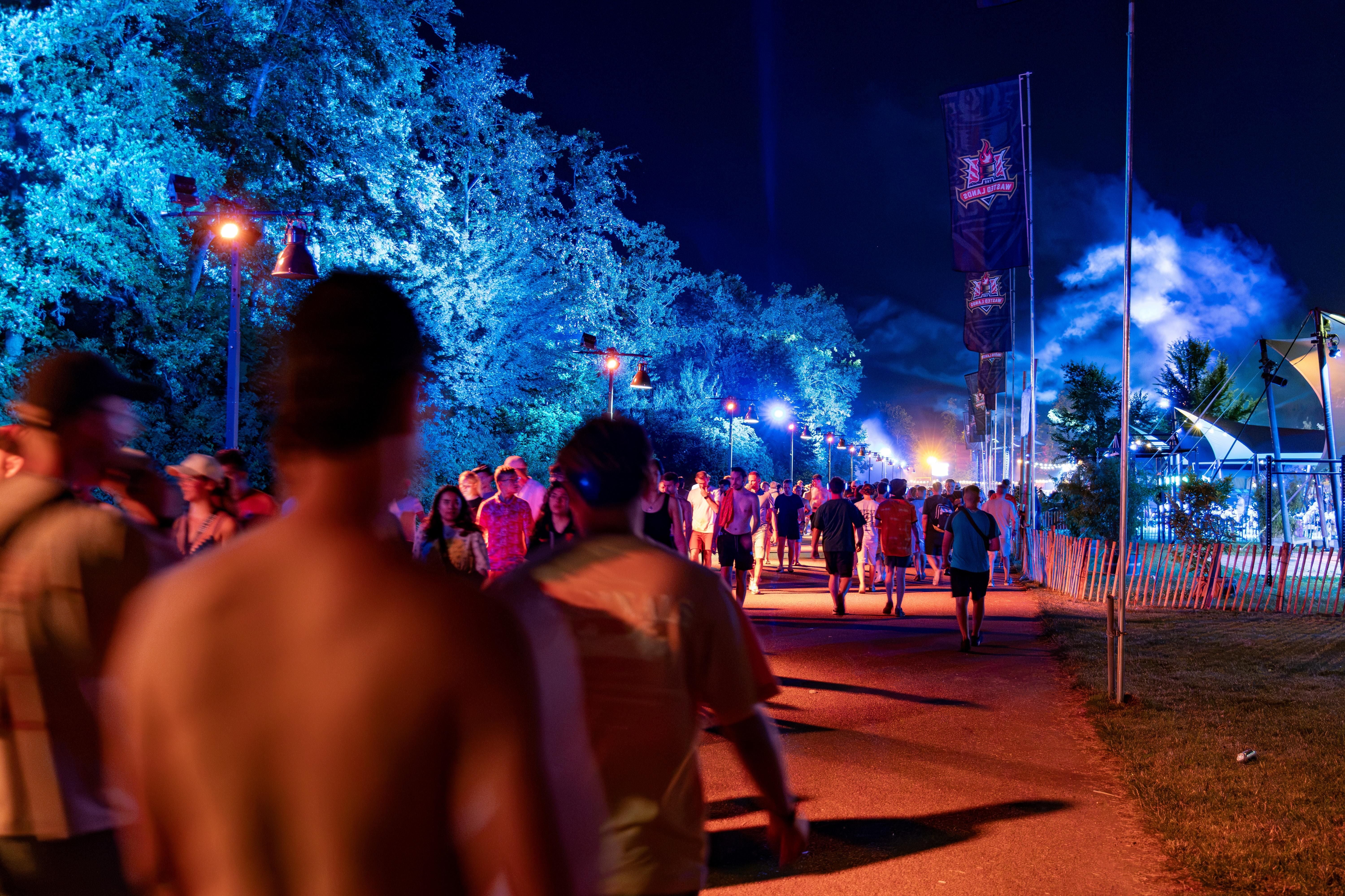 Menschen verlassen nachts ein beleuchtetes Festivalgelände, Bäume sind blau angestrahlt.