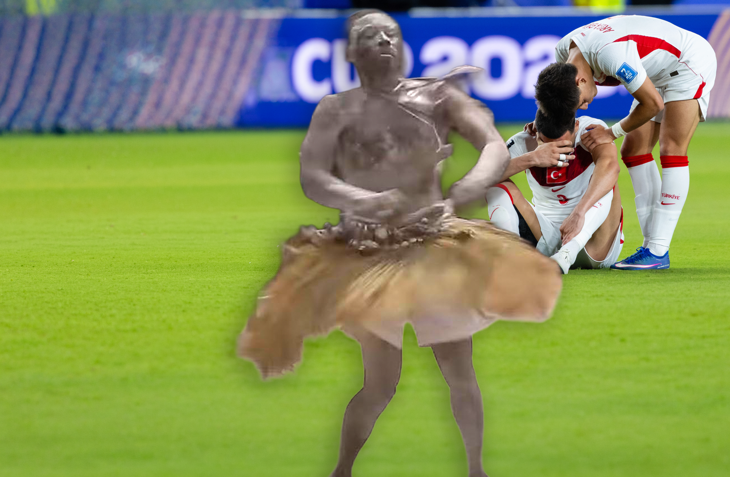 Fotomontage auf einem Fußballplatz. Im Vordergrund ist die halbtransparente, in Wischoptik freigestellte Figur eines traditionell gekleideten, tanzenden Mannes in einem Bastrock zu sehen. Im Hintergrund sitzen zwei enttäuschte Fußballspieler im weißen Nationaltrikot der Türkei auf dem Rasen; einer hält sich frustriert das Gesicht vor Augen, der andere beugt sich tröstend über seine Schulter.